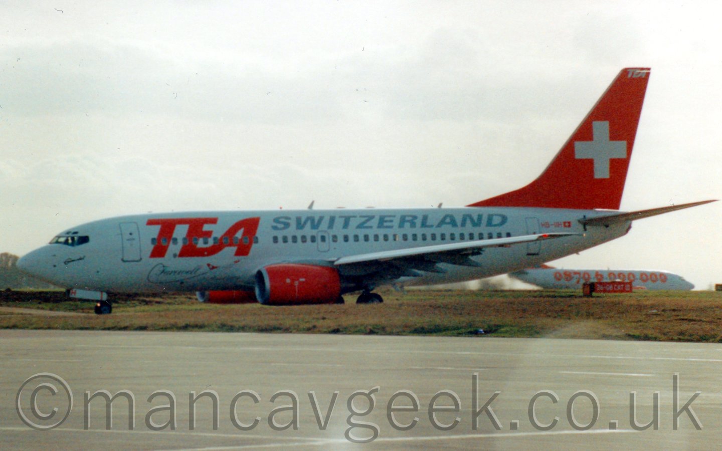 Side view of a twin engined jet airliner taxiing from right to left. The planes body is almost entirely white, with ted billboard-style "TEA" titles covering the cabin windows on the forward fuselage, with grey "Swizerland" titles above the windows on the rear fuselage. The engine pods and tail are red, with a large white cross on the tail. Large grassed areas run towards the camera in the foreground, and off into the distance in the background. Another white jet airliner with orange "0870 6 000 000" titles along the fuselage is visible in the background, just under the tail of this plane on the middle right of the frame. Above, the sky is a bright yet hazy grey.