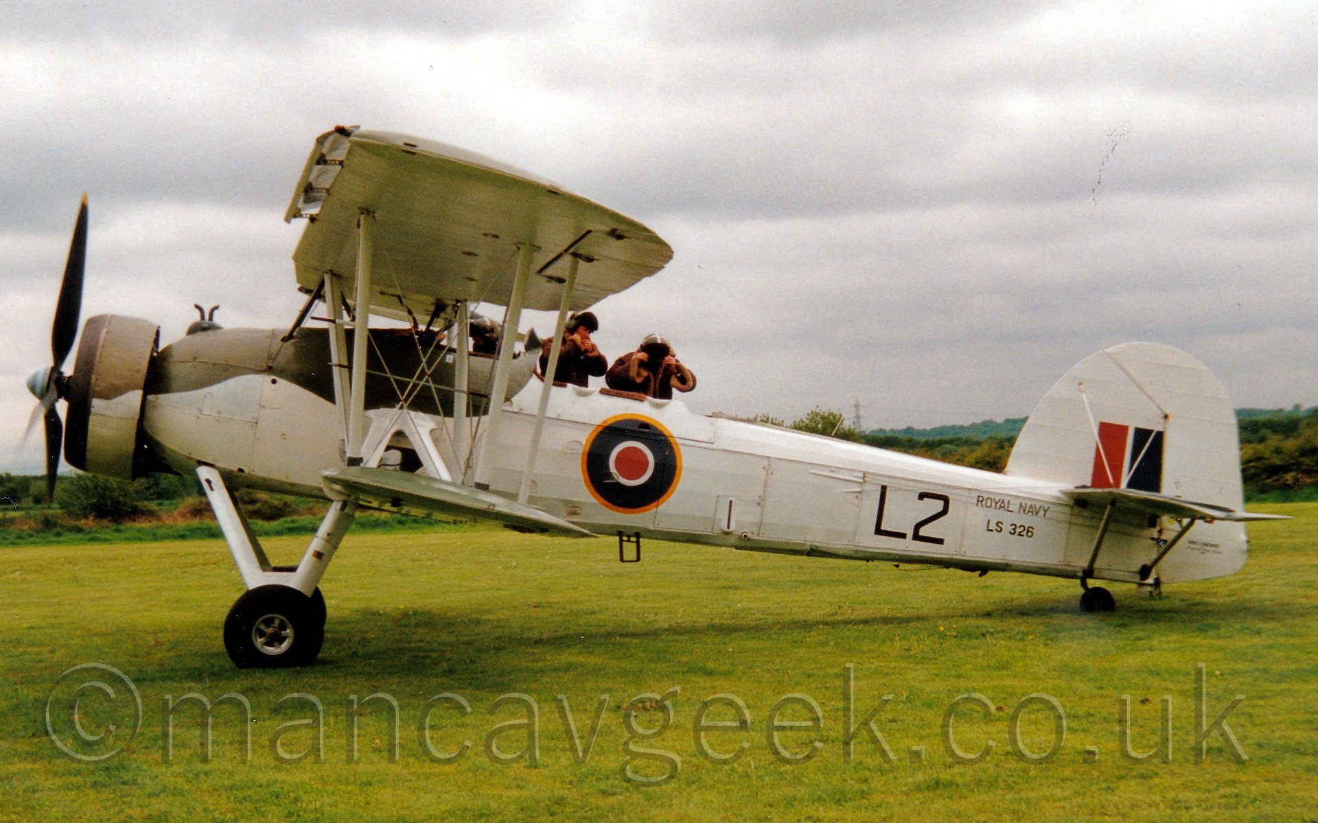 Side view of a World War II-era single engined biplane parked on grass facing to the left. The plane is mostly white, with an olive green section over the top of the nose and the engine cowling. There are 3 open cockpit positions, with the front one containing a helmeted pilot looking down at his instruments, the other two having helmeted crew wearing heavy fur coats standing and adjusting their helmets. below the rear cockpit is a yellow, blue, white, and red roundel. On the rear fuselage are large "L2" titles, with smaller "Royal Navy" and "LS326" titles further back. There are short, vertical red and blue bands on the tail. In the background, bushes mark the perimeter of the airfiled, sctretching off to the horizon, under a cloudy grey sky.