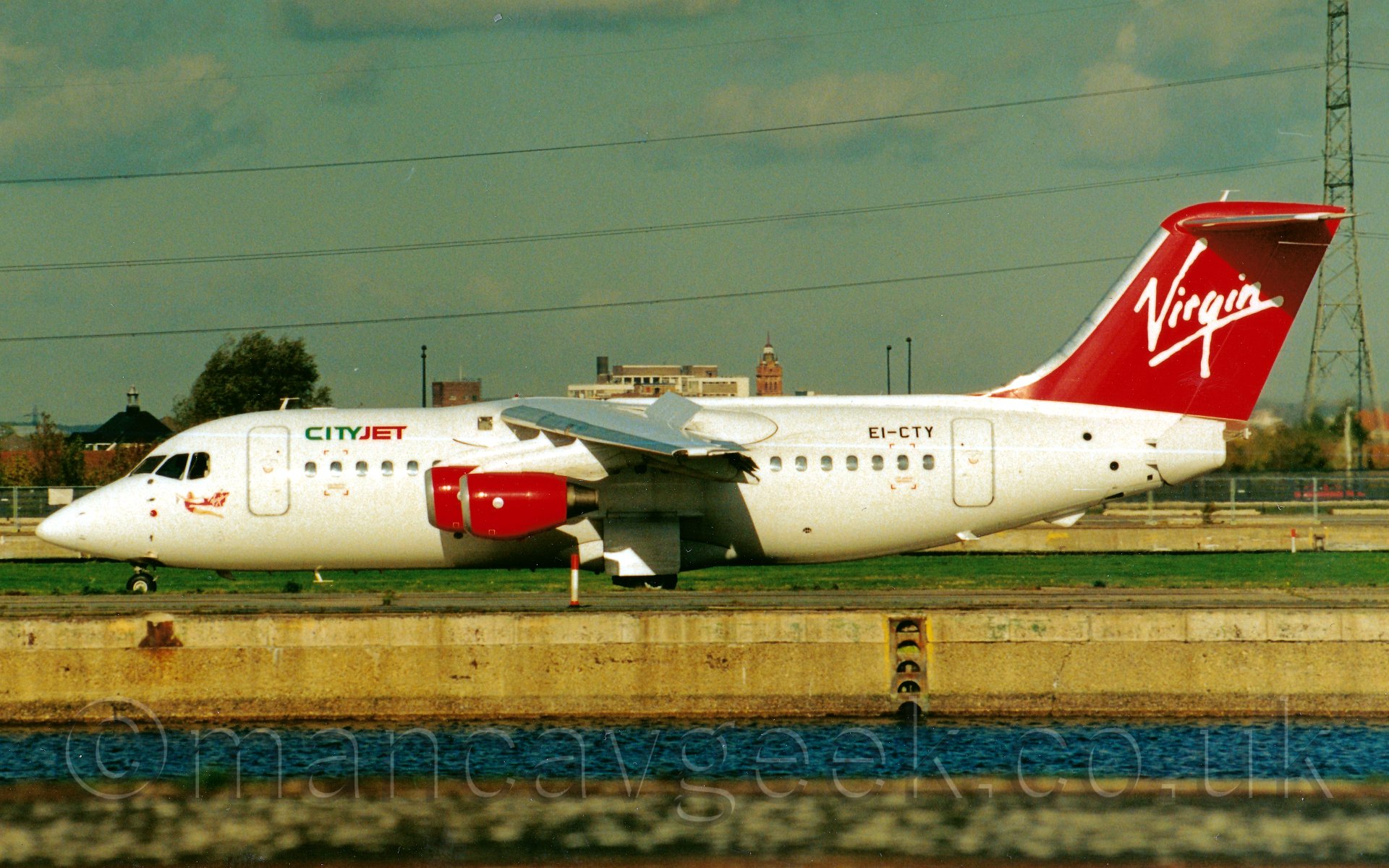 Side view of a high-winged, 4 engined jet airliner taxiing from right to left. The plane is mostly white, with green and red "CITYJET" titles on the upper forward fuselage, and an image of a flying woman weraing a red leotard and holding a red flag with the word "Virgin" written on it. The tail is red, with the word "Virgin" in the middle. The engine pods are also red. The registration "EI-CTY" is on the upper rear fuselage in black. In the foreground, a body of water can be seen, several metres below the level of the taxiway. In the background, green grass leads up to fences at the wirfield perimeter, with cars driving past on a nearby road, and houses and other buildings beyond that, under a blue-grey sky.