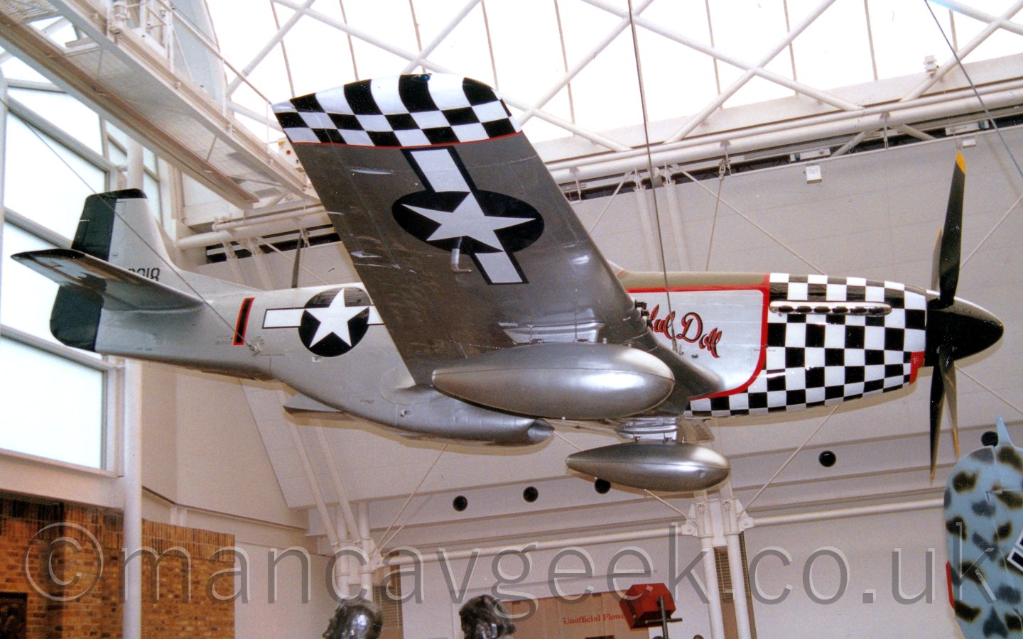 Side view of a World War II-era single engined fighter aircraft suspended from the roof of a museum, to replicate it's appearance in flight. the plane is mostly a silver colour, with a black and white checkerboard pattern over the engine cowling in the nose, and over the wingtips, outlined in red. There are white circles containing a white, 5-pointed star, and a white bar outlined in blue on each side, on the rear fuselage and under the wings. In the background, the white walls and curved glazed roof of the museum provide great, flat lighting for the room.
