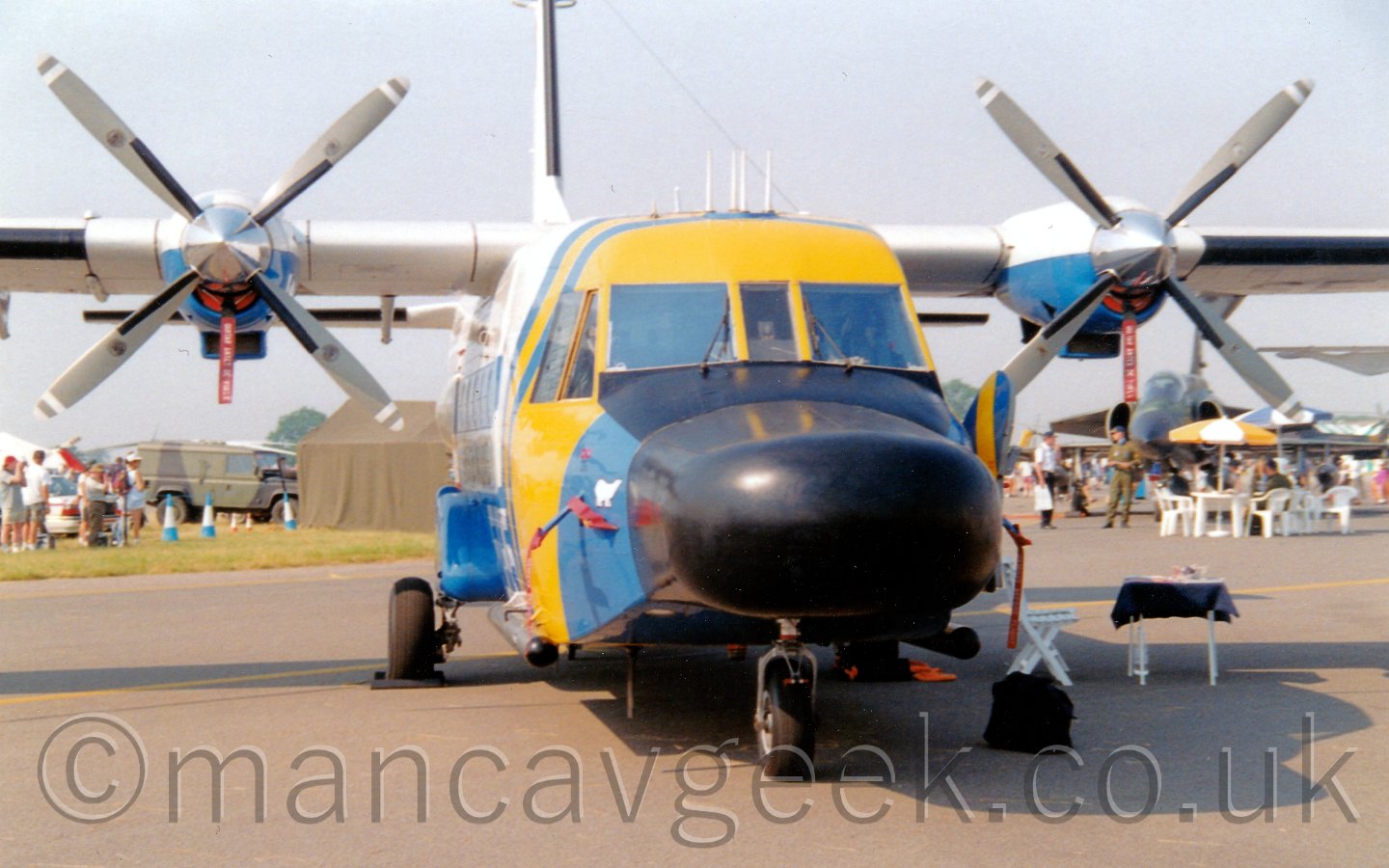 Front view of a high-winged, twin propellor engined transport aircraft, facing slightly to the right of the camera. The plane plane is mostly white, with blue and yellow bands around the forward fuselage. The nose has a big radome, painted black, looking a bit like a duck's beak. There are pods on the side of the lower rear fuselage for the main landing gear. In the baclkground, small crowds of people are walking around, looking at some of the other exhibits, including a jet fighter aircraft under this plane's wing, under a pale grey sky.