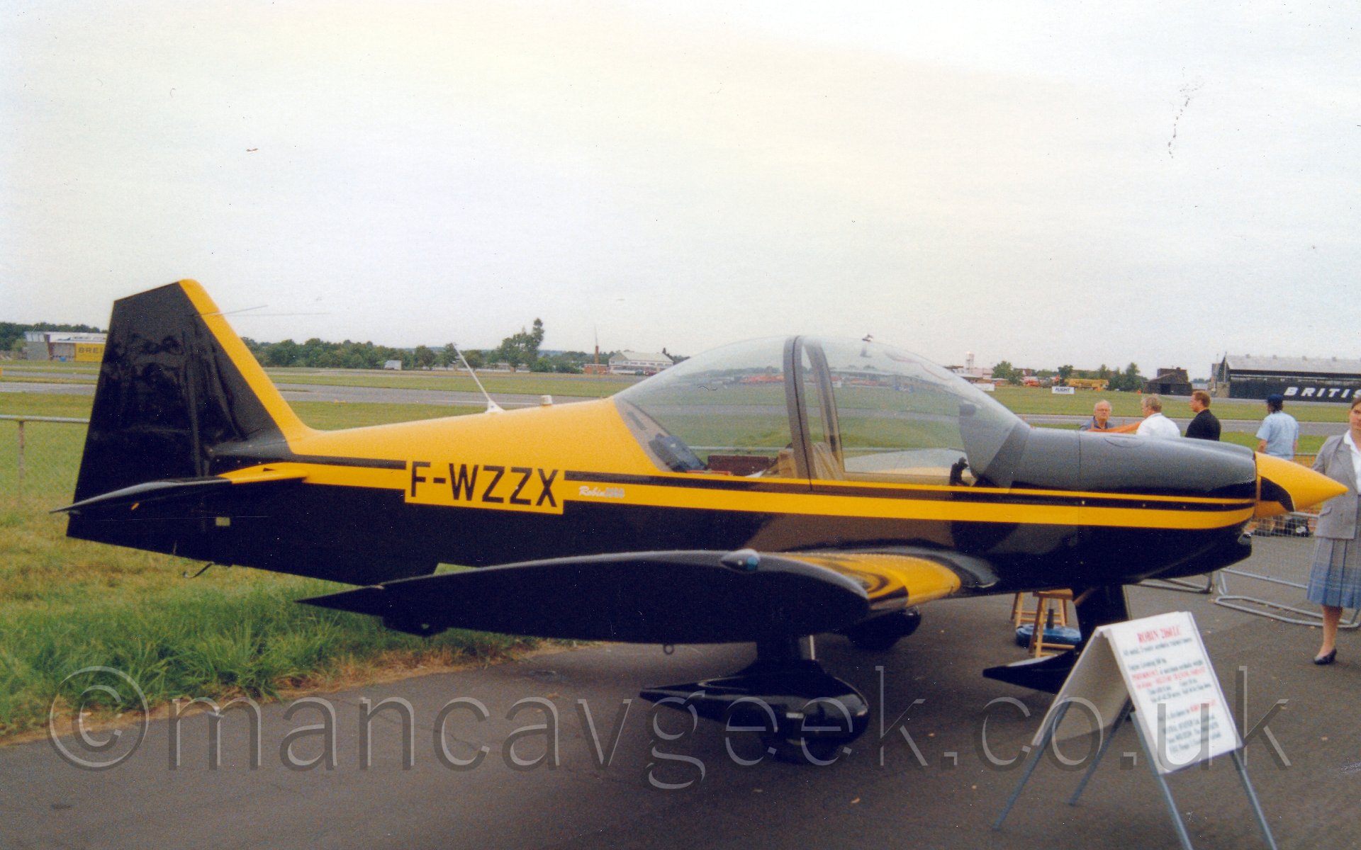 Side view of a single-engined, 2 seat light aircraft parked facing to the right. The plane is mostly black, with with a yellow stripe runnning along the body and the front edge of the tail, with the top of the rear fuselage aft of the cockpit also being yellow. The plane has a fixed undercarriage, with black spats over the wheels. There is a white A-frame holding some information about the plane for anyone who may be interested. Some people can be seen standing around on the far side, behind a waist-level metal fence. In the background, a large black hangar can be seen on the far side of the airfield on the left of the frame, with other buildings and trees stretched across the rest of the frame, under a cloudy grey sky.