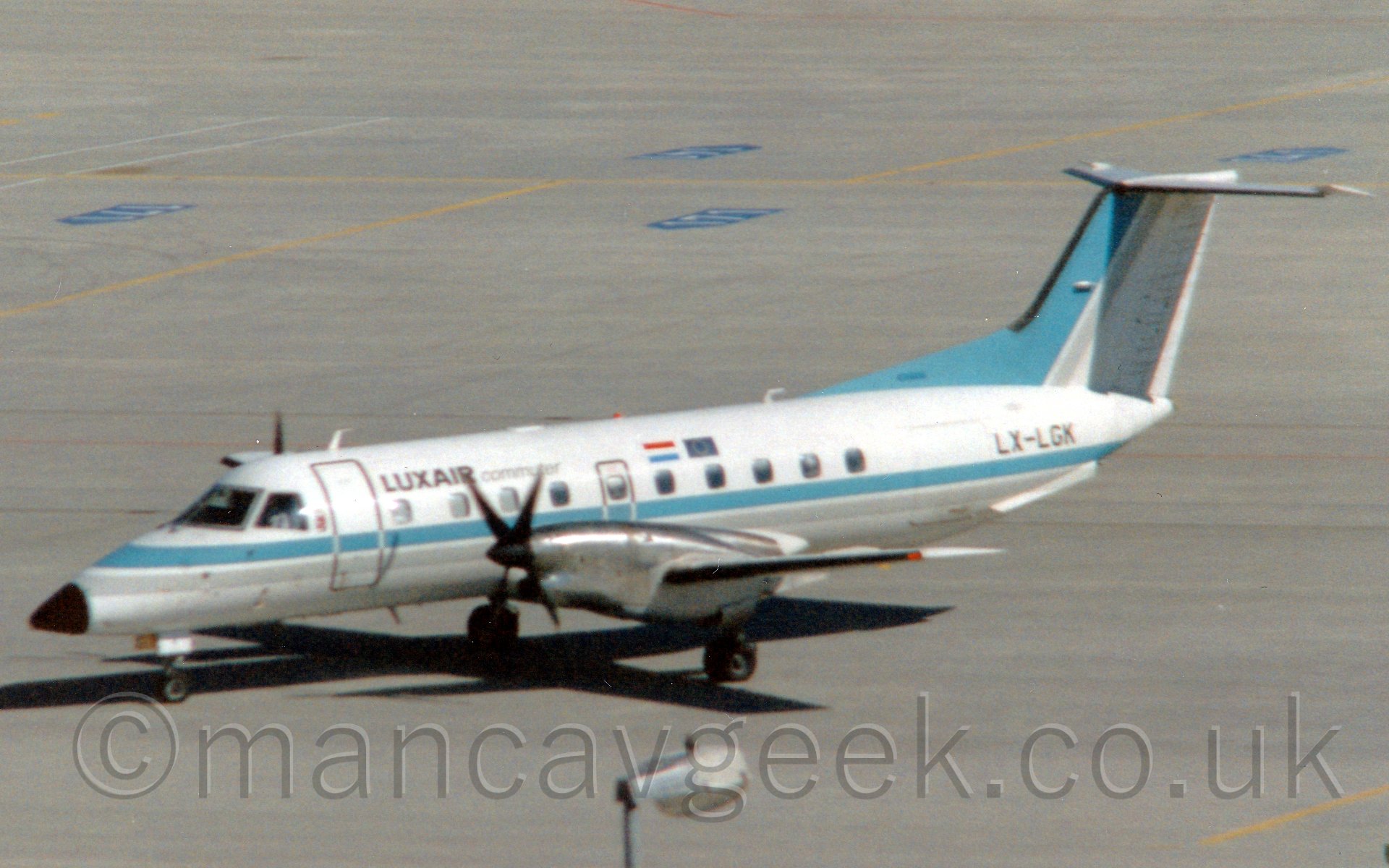 Side view of a twin propellor-engined airlinertaxiing from right to left. The plane is mostly white, with a thick light blue and light grey stripe running along the body from the nose. There are small "Luxair" titles on the upper forward fuselage, just aft of the forward door. The tail is mostly white, with a light blue leading edge, and light grey diagonal stripe. The engine pods are a bare metal finish. In the background, acres of concrete fill the screen.