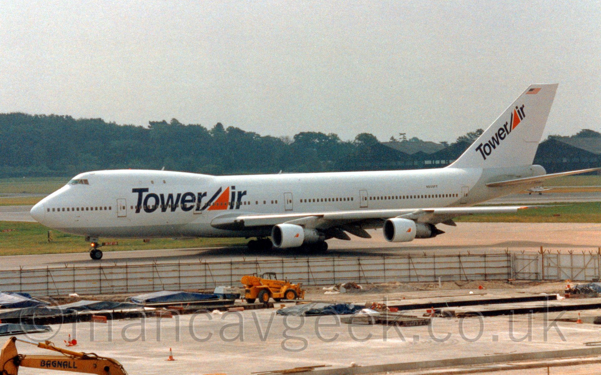 Side view of a very large 4 engined jet airliner taxiing from left to right. The plane is mostly white, with a silver belly, and large "TowerAir" titles on the forward fuselage, with the centre of the "A" in "Air" being a large orange triangle. The same titles are painted diagonally on the tail. In the foreground, a large area is fenced off, with several yellow constructions vehicles scattered around, on what looks like newly poured areas of concrete. In the background, a large black hangar is on the left of the frame behind the tail, while trees fill the skyline on the rest of the frame. Above, the sky is a bright, hazy grey.