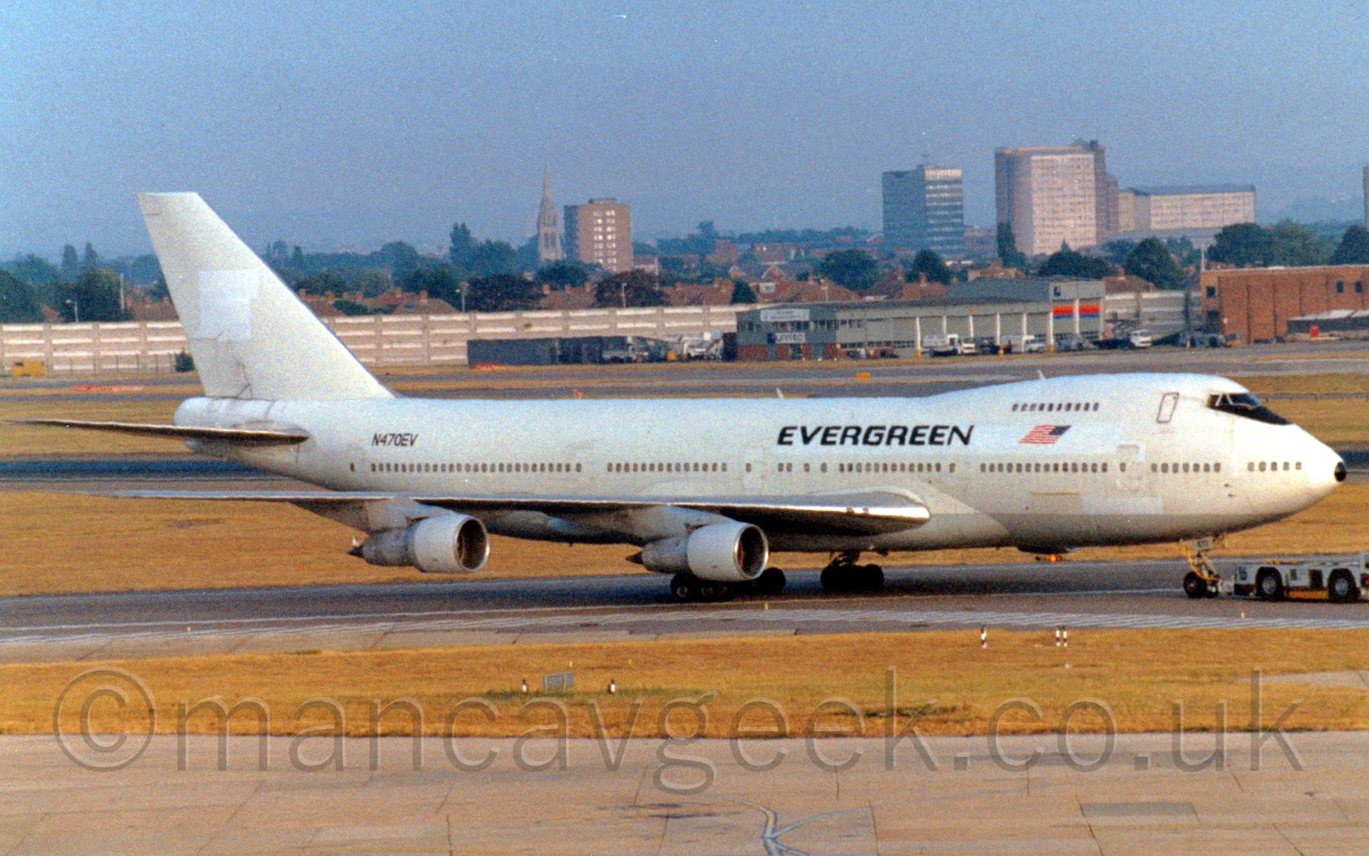 Side view of a very large 4 engined passenger airliner being towed from left to right on a taxiway by a dark blue and grey tug. The plane is mostly white, with the top of the nose cone painted black, a reversed US flag between the upper and lower deck windows on the forward fuselage, and large black "Evergreen" titles over the front of the wings. The registration "N470EV" is painted prominently on the upper rear fuselage. In the background, a grey building is right up against a large concrete retaining wall, with houses, a church, and some tower blocks behind it, under a blue-grey sky.