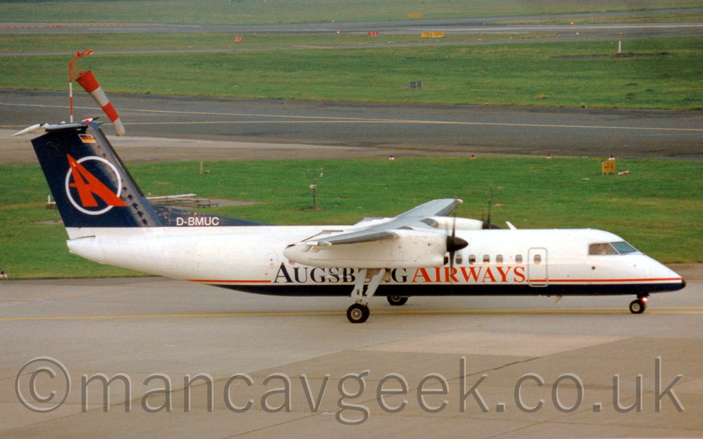 Side view of a high-winged, twin propellor-engined airliner taxiing from left to right. The plane is mostly white, with a dark blue belly, with a thin red stripe runniing along the body seperating the two colours. There are dark blue and red "Augsburg Airways" titles on the lower fuselage. The tail is dark blue, with the white outline of a circle, overlaid with a red capital "A", pointing to the North-West, and vaguely resembling a runway. In the background, grassed areas are seperated by taxiways and a runway.