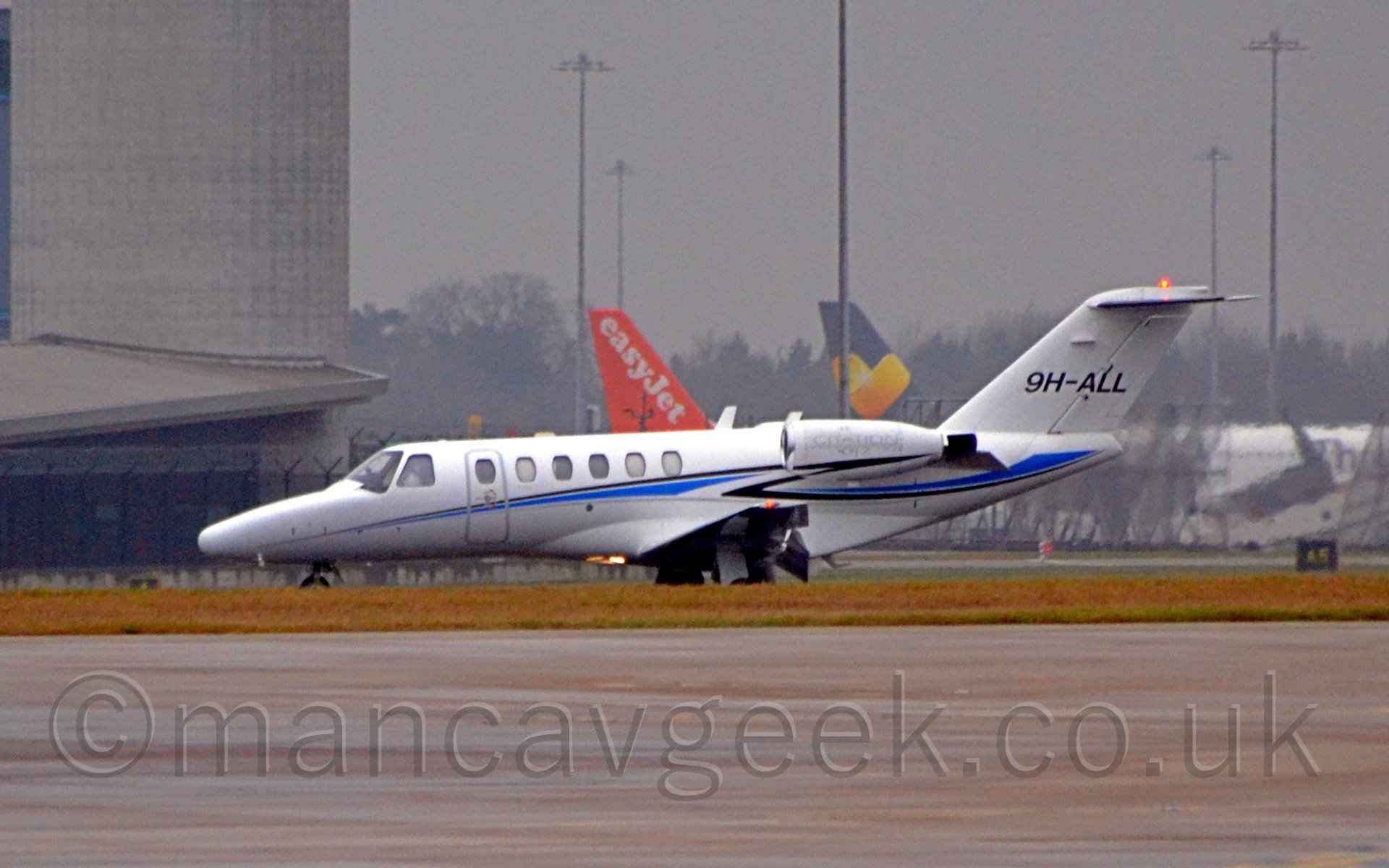 Side view of a twin engined bizjet moving from right to left after landing on a runway. The plane is mostly white, with a blue and black stripe running along the fuselage and under the engines. The registration "9H-ALL" is displayed prominently on the tail, with additional grey "CITATION" titles on the engine pods. The flaps are deployed from behind the wings. In the foreground, a wide stretch of tarmac leads up to a gressed area, with the grass partially obscuring the planes wheels. In the background, a tall grey tower dominates the left of the frame, while orange and grey tails are visible behind this plane in the distance, with scattered lighting poles stretching into the moody grey sky.