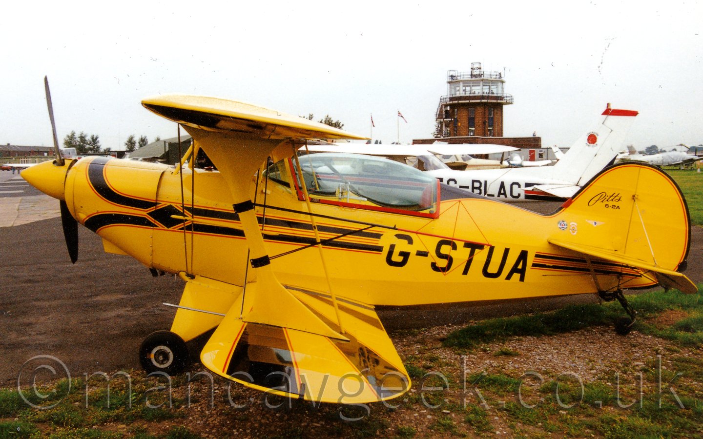 Side view of a single engined, single seat biplane parked facing to the left. The plane is mostly yellow, with a pair of black stripes outlined in red running along the body, overlaid on the rear fuselage with the registration "G-STUA". The wings are staggered, the top one being quite a bit further forward than the bottom one. In the background, a selection of other light aircraft are parked, with a brown tower with a fully glazed room at the top in the distance. The sky above and behind everything is a hazy grey.
