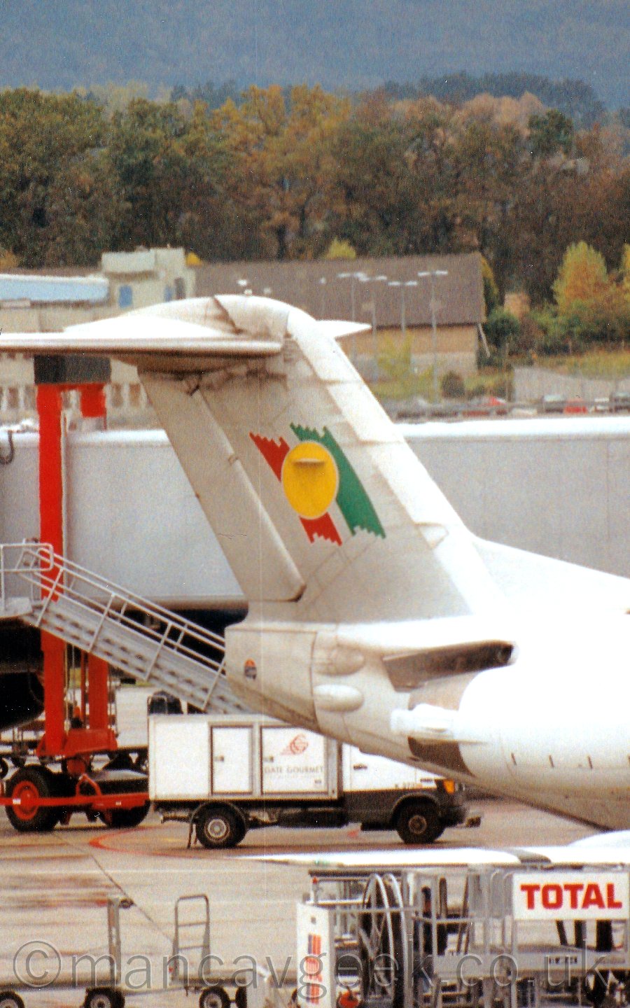 Close up of the rear fuselage and tail of a twin engined jet airliner parked facing to the right. The plane's body is white, while the tail is a light grey, with short red and greendiagonal bands in the middle, overlaid with a yellow circle. In the backgrouund, a white vehicle can be seen under the tail, while an airbridge stretches all the way across the middle of the frame. In the distance, a brown building stands in front of a load of trees, with a tree-clad hill climbing to the top of the frame.