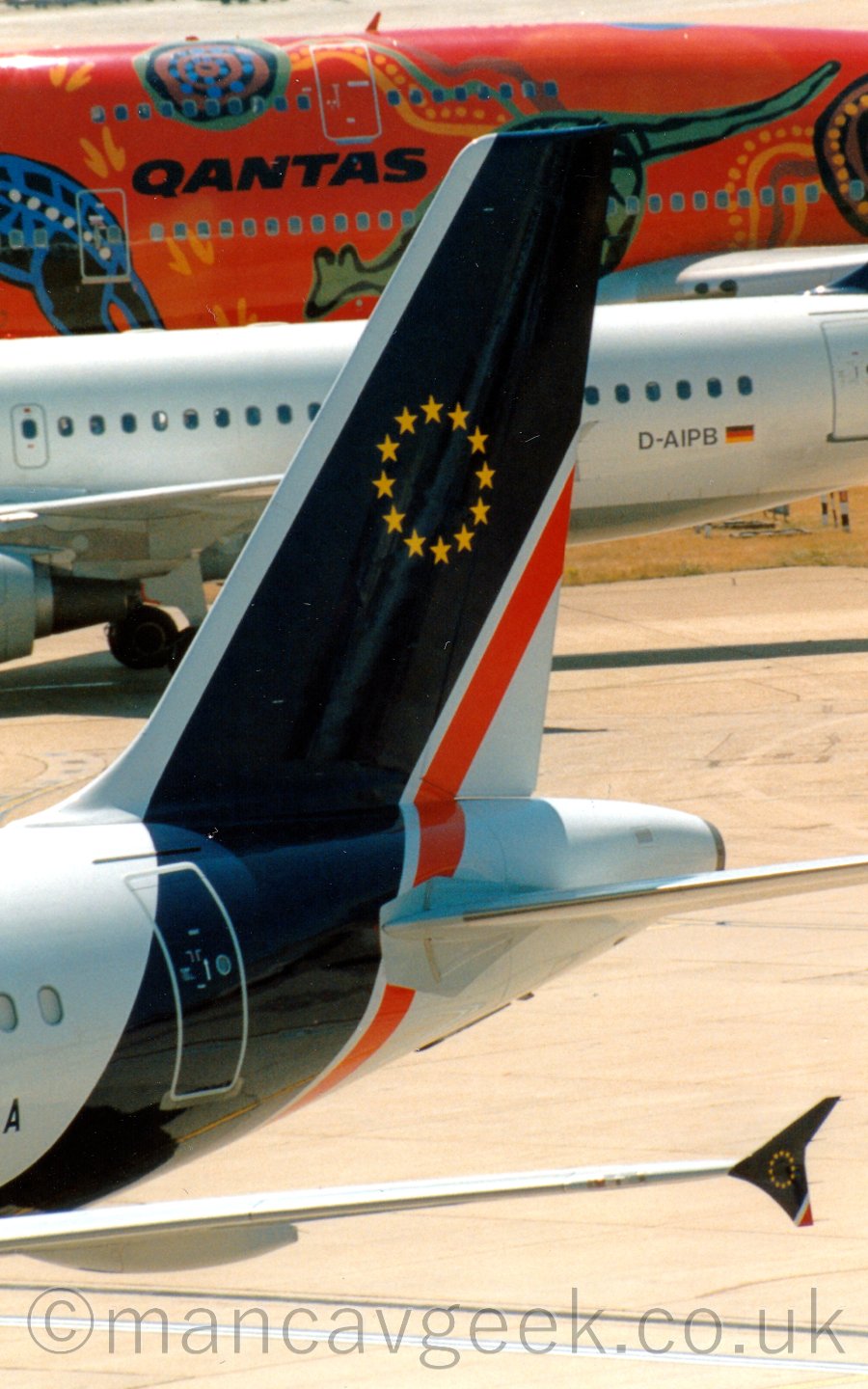 Closeup of the tail of a twin engined jet airliner parked facing to the left. the planes body is mostly white, with a thick dark blue and thin red band running round the rear fuselage and up in to the tail, where there is a circle of 12 5-pointed gold stars. The same design is visible on the planes upturned wingtips, at the bottom of the frame. Strong sunlight is casting strong shadows, and causing bright reflections on the upper surfaces. In the background, a white jet airliner and another, much larger, orange jet airliner are passing through the image.