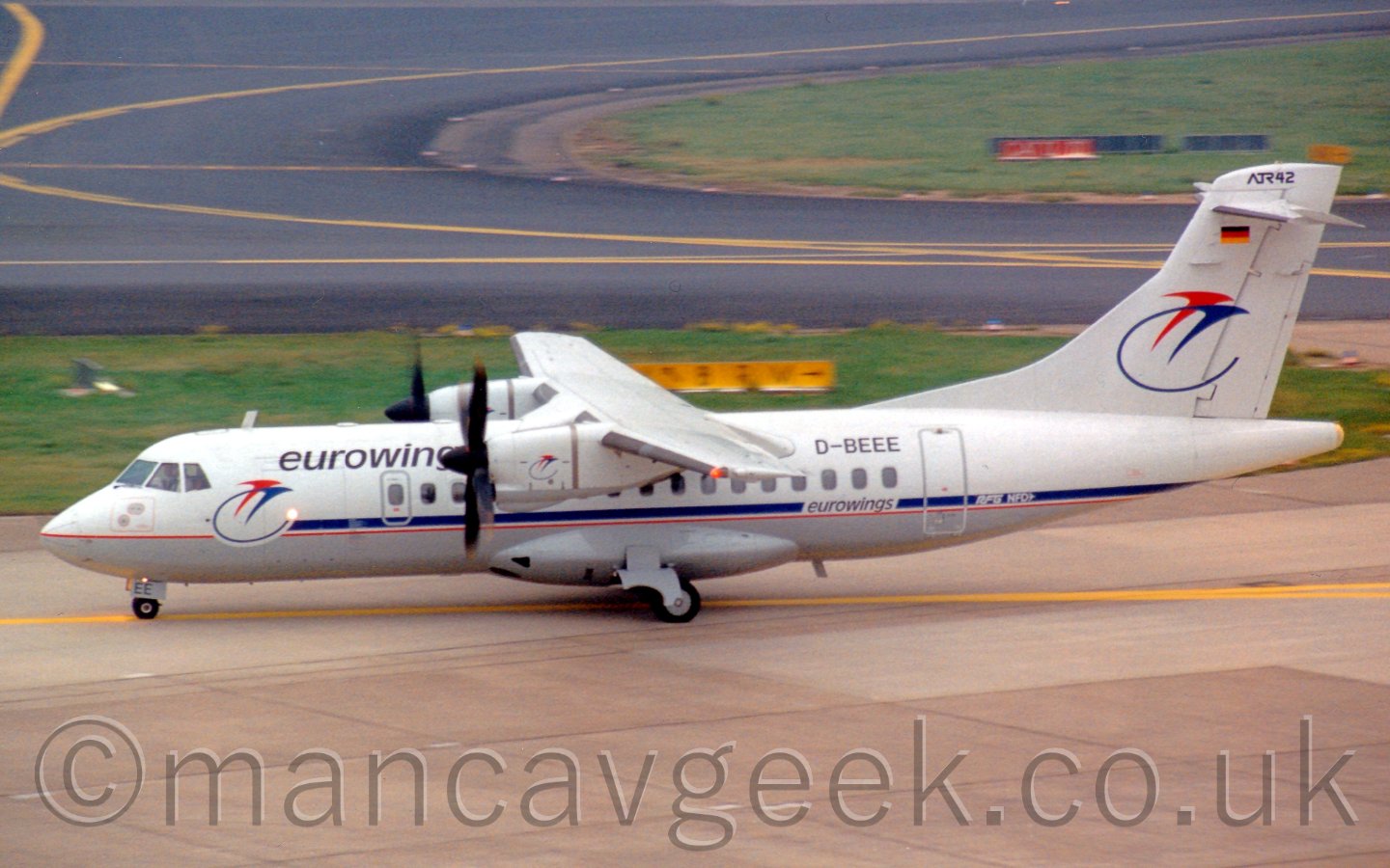Side view of a high-winged, twin propellor-engined airliner taxiing from right to left. The plane is mostly white, with a grey belly, seperated by a red and blue stripe along the body. There are "eurowings" titles on the upper forward fuselage, slightly above and aft of a stylised image of two flying birdss, one red, one blue, with the blue one having a tail that loops below in a circle, this same logo appearing in a larger form on the tail. The registration "D-BEEE" appears of the upper rear fuselage, with small "eurowings" titles below that. In the background, taxiways bisect large grassed areas.