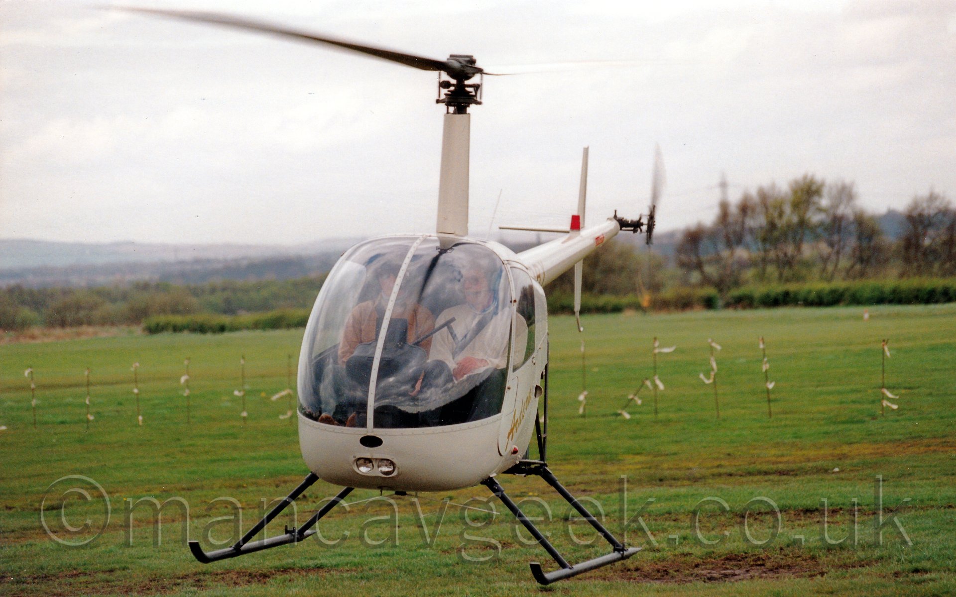 Front view of a small helicopter hovering just inches above the ground. The body, tail boom, and rotor mast are mostly white, with some indeciherable golden writing on the doors. Two people can be seen through the large cockpit windows. In the backgroud, the grass airfield stretches off into the distance, it's perimeter marked by bushes and trees. Metal poles are planted in the ground, with white markers fluttering in the breeze. Rolling hills in the far distance merge with the hazy grey skies.