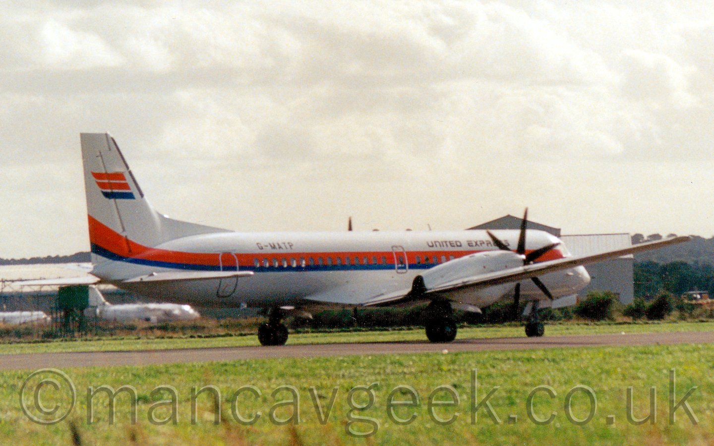 Side view of a twin propellor-engined airliner taxiing from left to right and slightly away from the camera. The plane is mostly white, with a red, orange, and blue stripe running along the body, covering the passenger cabin windows. There are black "United Express" titles on the upper forward fuselage, with the registration "G-MATP" on the upper rear fuselage. The tail has 3 red, oroange, and blue horizontal bands. In the background,a low, grey hangar stretches across the frame, with a couple of white planes parked in front of it on the middle left, under a bright but hazy sky.