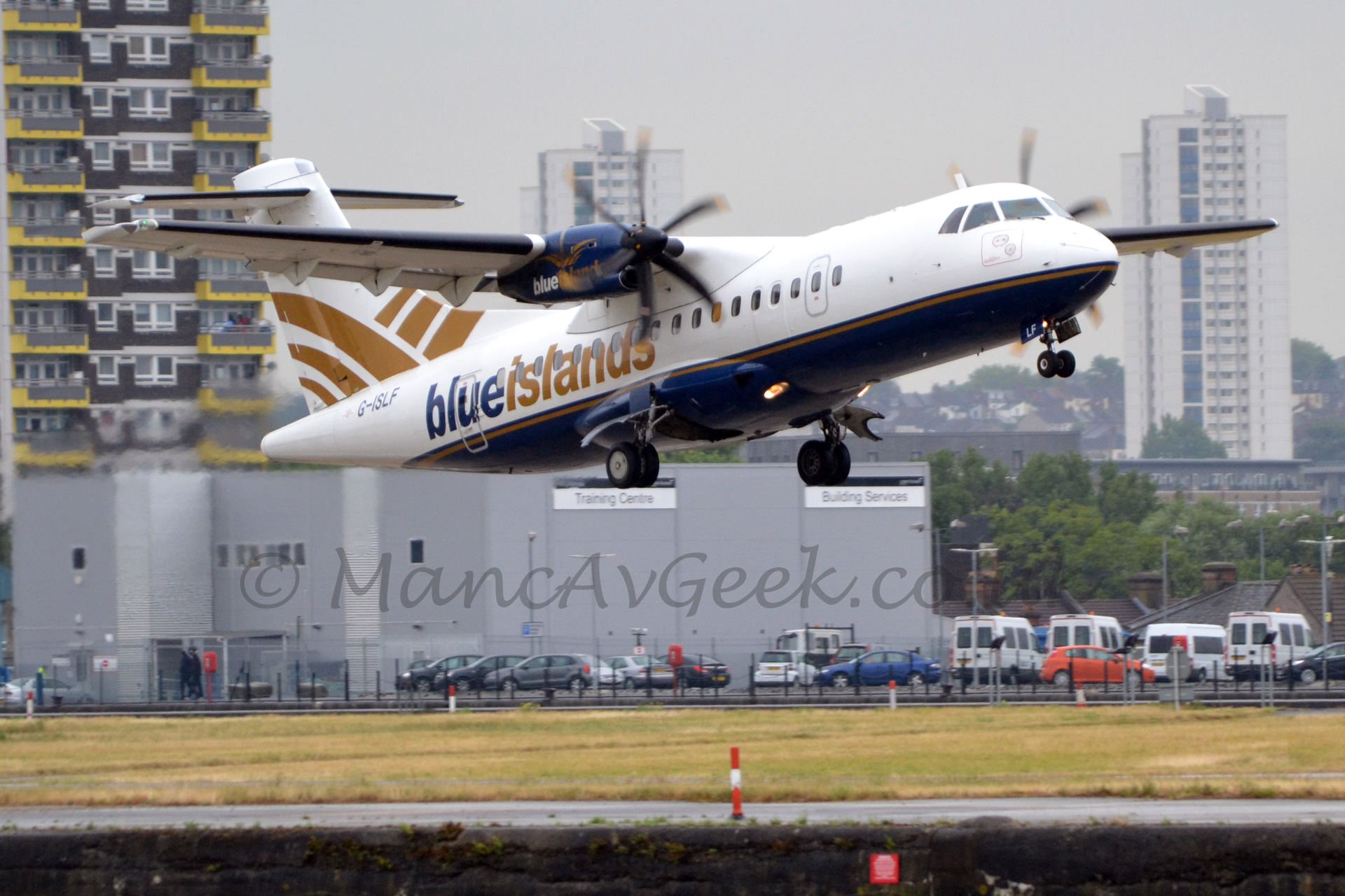 Side view of a high-winged, twin propellor-engined airliner flying from left to right at a very low altitude with it's undercarriage lowered and flaps extended from behind the wings, and the nose raised sharply, suggesting it has just taken off.. The plane is mostly white, with a blue belly, and a golden stripe running along the body. There are large blue and gold "blue islands" titles on the rear fuselage, with the registration "G-ISLF" under the tail. The engine pods are blue, with white and gold "blue islands" titles. The tail is white, with some ciurved golden stripes. In the background, grass stretches off in to the distance, with some cars parked in front of a large grey building beyond that. Hi-rise tower blocks dominate the skyline, under a dreary grey sky.