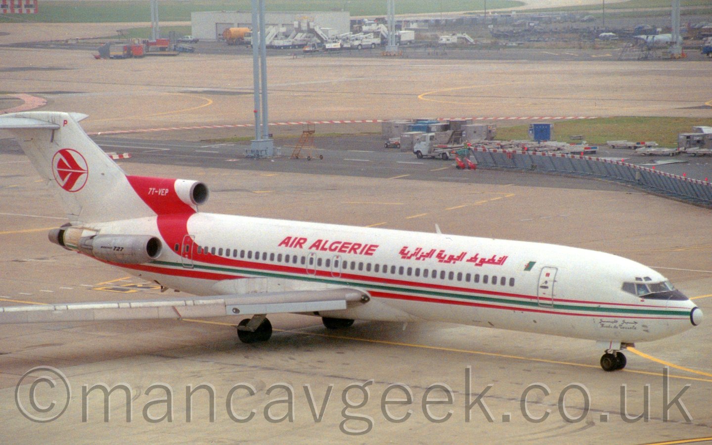 Side view of a 3 engined jet airliner with the engines mounted on the rear fuselage. The plane is mostly white, with a red/green/red stripe running along the body, the upper red part sweeping up over the tail-mounted engine, with the registration "7T-VEP". There are red "AIR ALGERIE" titles on the upper fuselage, in English above the wings, and in an Arabic script further forward. The side-mounted engines have a natural metal finish, with black "Boeing 727" titles. The tail is white, with the red outline of a circle, with the stylised image of a red flying bird in the middle. In the background, a grey metal barrier with alternating white and rad bands on the top runs from the middle left of the frame towards the centre, with some baggage carts parked behind it, with another parking area for vehicles at the top of the frame, next to a low grey building.