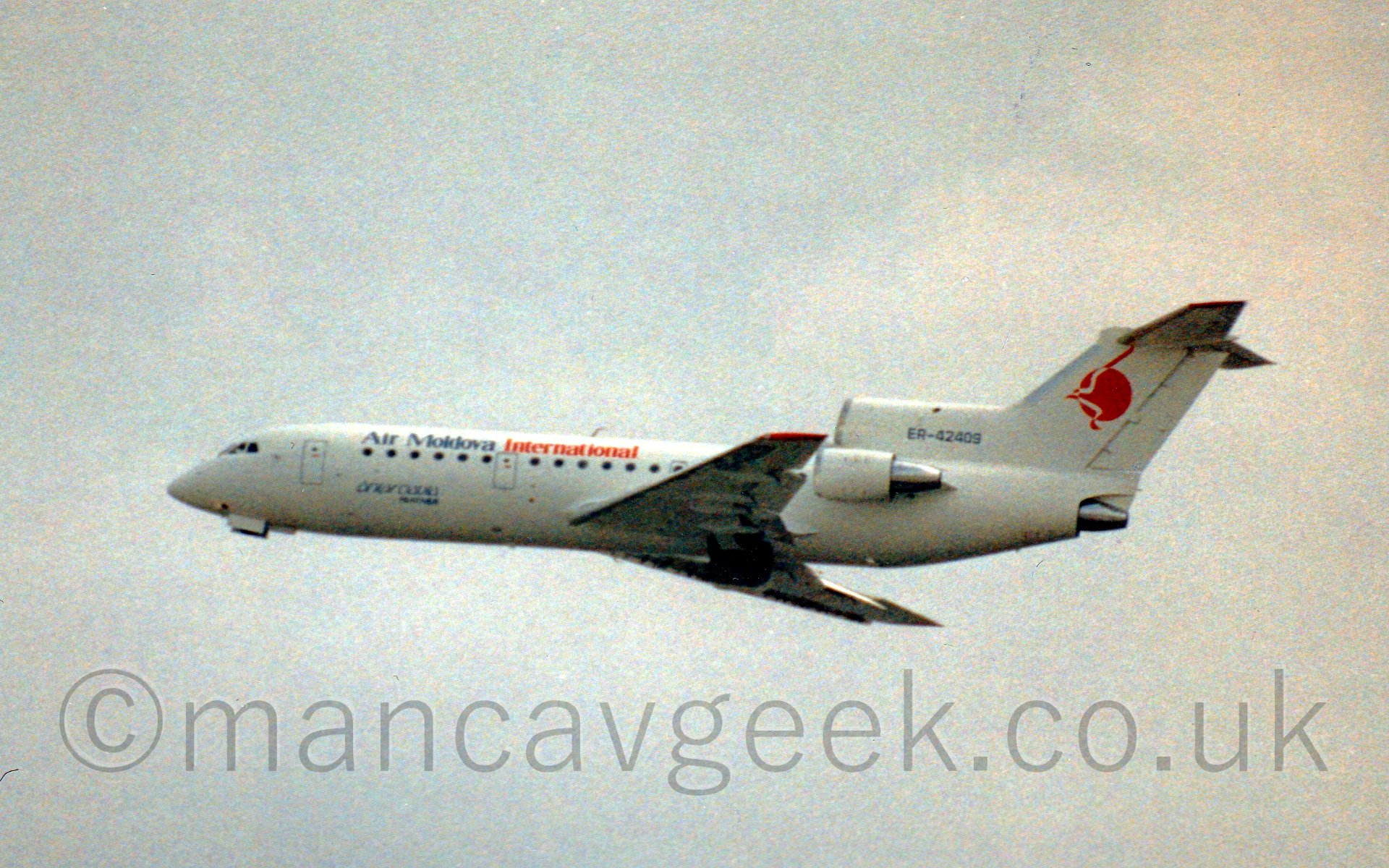 Somewhat grainy, hazy side view of a 3 engined jet airliner with the engines mounted on the rear fuselage, flying from right to left at a low altitude, with gear retracting, flaps deployed from behind the wing, and the nose raised, suggesting it has just taken off. The plane is almost entirely white, with blue and red "Air Moldova International" titles on the forward upper fuselage, and the registration "ER-42409" on the centre engine intake. The tail has a red circle, with a stylised white and red image of a flying bird overlaid. A hazy grey sky fills the rest of the frame.