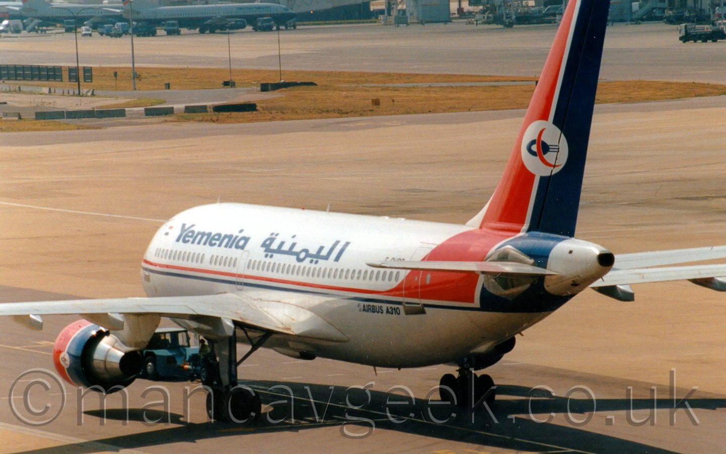 Side view of a twin engined jet airliner being pushed back from it's stand, facing to the left and away from the camera. The plane is mostly white, with a thin red and blue stripe running along the body and sweeping up to fill the tail.There are dark blue "Yemenia" titles on the upper fuselage, in English at the front, and in an Arabiv script towards the middle. The tail has a white circle, with a dark blue oval with a white hoile, and 3 lines trailing off the back, with a red crescent overlaid on top. There are "Airbus A310" titles on the lower rear fuselage. The engine pods are red at the front, and blue at the rear. There is a blue tow truck attached to the nose gear. In the backgrpound is a view across the airfield, mostly concrete apron, with a handful of airliners parked by a large building in the top left corner.