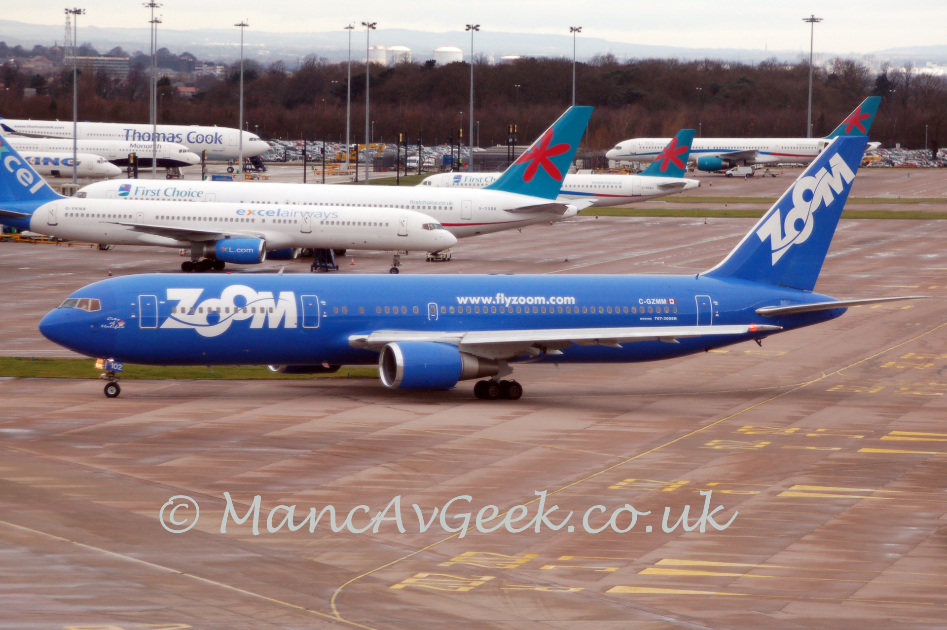 Side view of a twin engined jet airliner taxiing from right to left. The plane is almost entirely blue, with large white billboard-style "Zoom" titles on the forward fuselage, and smaller "www.flyzoom.com" titles on the upper fuselage over the wing, and the registration "C-GZMM" on the upper rear fuselage, next to a Canadian flag (red stripes either side of a white field, with a red maple leaf in the centre). There are large white diagonal "Zoom" titles on the tail. In the background, an array of twin engined airliners are scattered around, nestled among tall lighting poles, with tree marking the edge of the airfield, and an industrial landscape beyond that, under a hazy grey sky.
