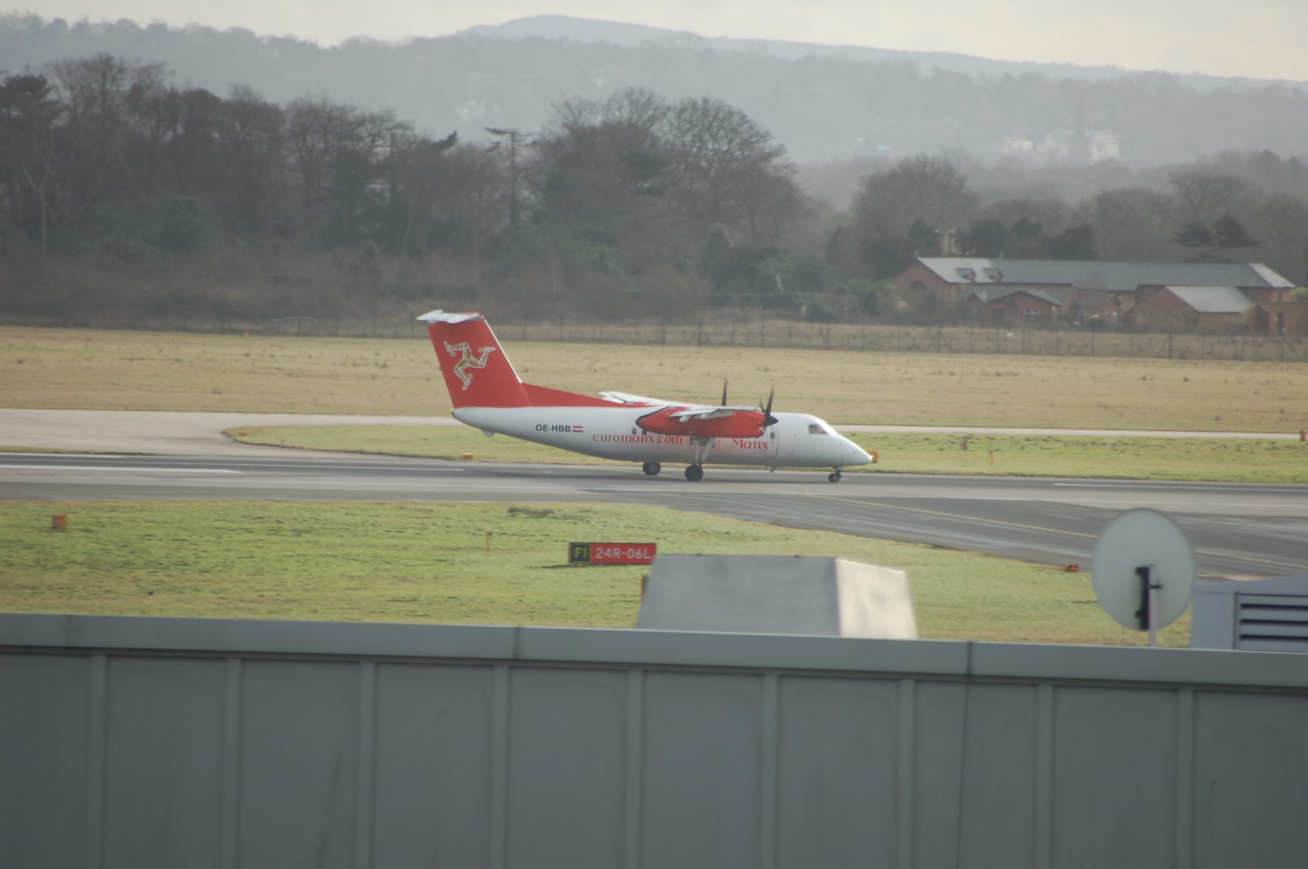 Side view of a high-winged, twin propellor engined airliner taxiing from left to right after landing on a runway. The plane is mostly white, with red "euromanx.com" on the lower rear fuselage, and "Manx" titles on the lower forward fuselage. The registration "OE-HBB" is on the rear fuselage in black, next to an Austrian flag (equal red-white-red horizontal bands). The tail and engine pods are red, with 3 goldenn, armoured legs, joined at the hip and running in a circle on the tail. In the foreground, the lower quarter of the frame is a grey building, with grass beyond that running to the runway. In the background, large grassed areas lead off to trees at the airfield perimeter. On the middle right of the frame, a large brown brick building with a sloping black roof is visible. Rolling hills beyond that slowly vanish in to the hazy grey sky.