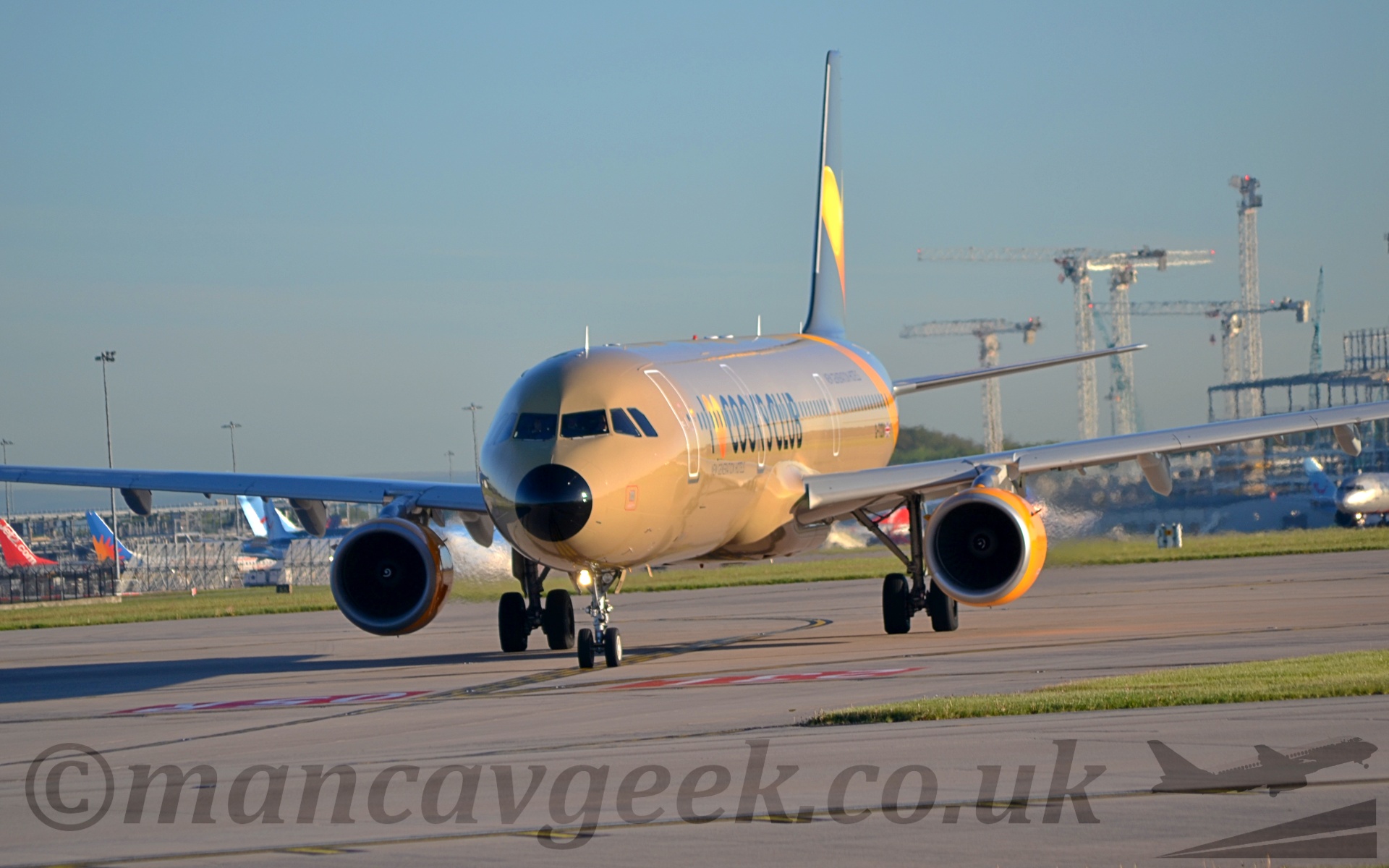 Side view of a twin engined jet airliner taxiing from right to left and slightly towards the camera. The planes body is an unfortunate shade of brown, with "I (heart) Cook's Club" titles on the forward fuselage, with a large yellow and orange heart shape in place of the word. The rear fuselage and tail are dark grey, with a diagonal yellow band running around the fuselage. The engine pods are the same bright yellow. In the background, several planes can be seen clustered under the left wing, while several tall cranes soar into the glorious blue sky above the left wing.