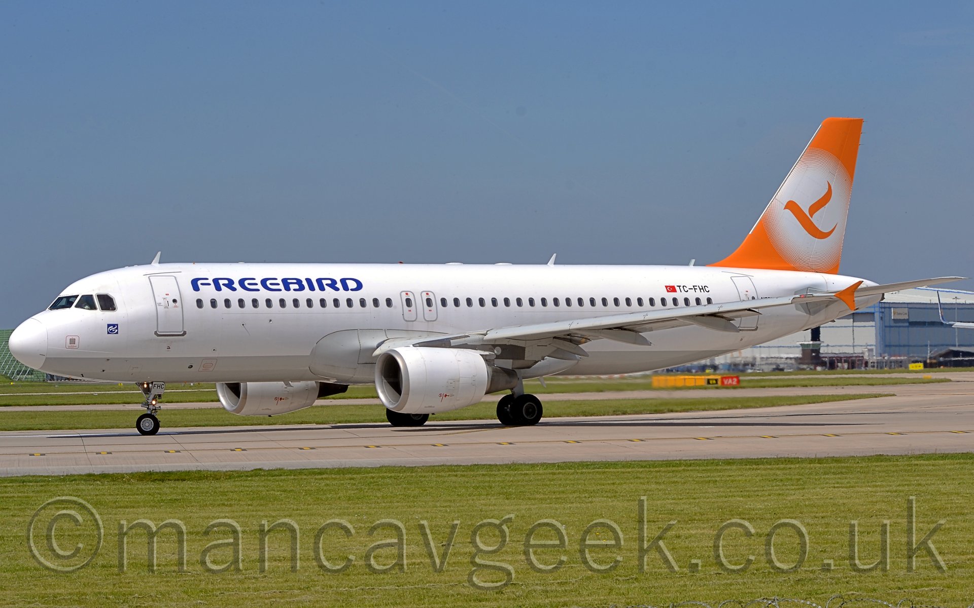 Side view of a twin engoinned jet airliner taxiing from right to left, turning slightly away from the camera. The plane is mostly white, with a grey belly, and blue "FREEBIRD" titles on the upper forward fuselage. There are orange vertical endplates on the wingtips. The tail is orange, with a white circle containing an orange flying bird. green grass fills most of the foreground, as well as a lare bit of background visible under the planes body. There is a large grey hangaron the laft of the frame, behind the planes tail. Above, the sky is a pale blue.