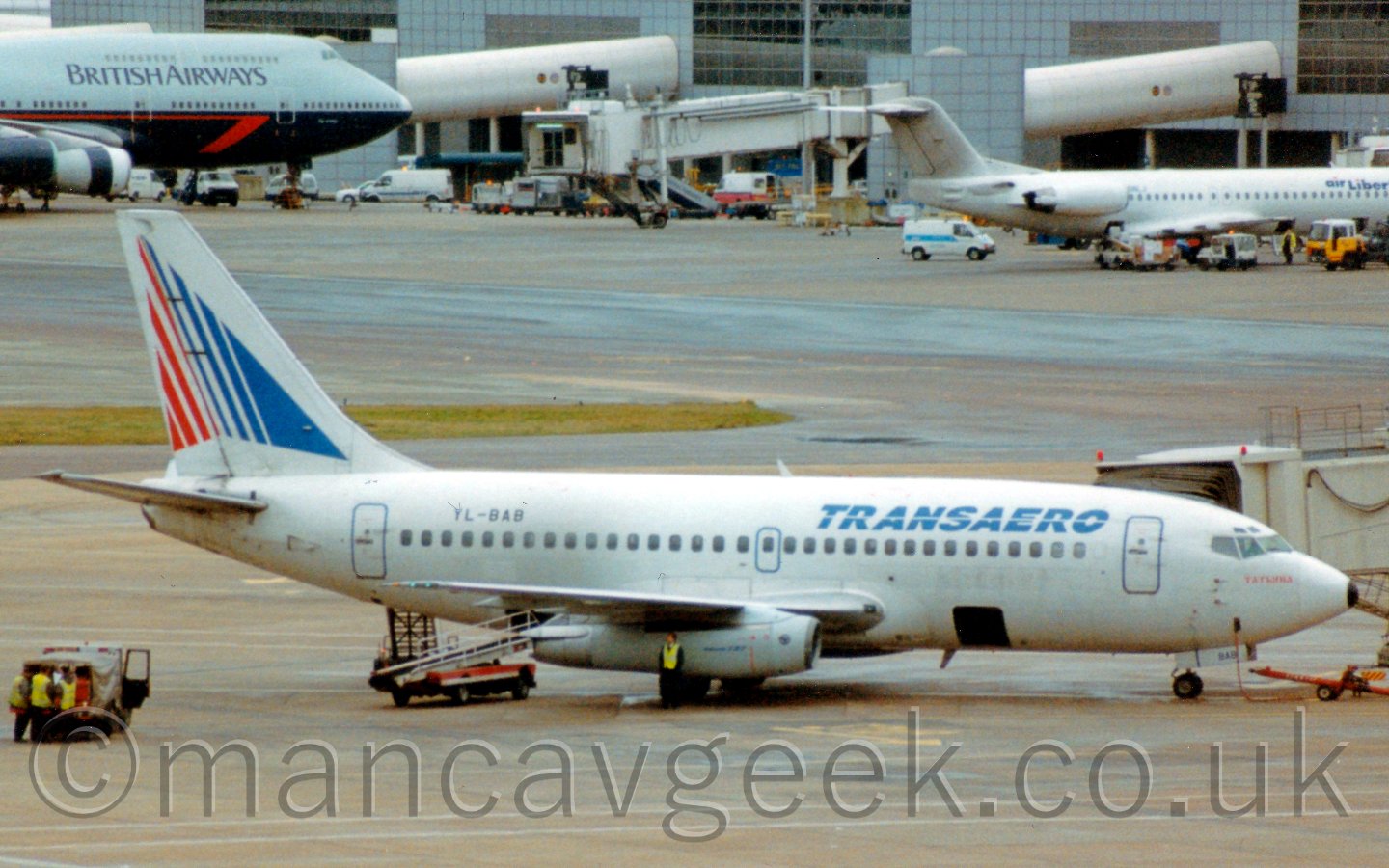 Side view of a twin engined jet airliner parked at it's gate, facing to the right, with a white airbridge attached to the forward door on the opposite side. The plane is mostly white, with blue "TRANSAERO" titles on the upper forward fuselage, and the registration "YL-BAB" on the upper rear fuselage. The tail has a large arrowhead design, pointing upwards, made up of vertical red and blue lines of varying widths. There are cargo bay doors open in the lower front and rear fuselagge, the one at the rear has an orange and white belt loader raised towards it. Some baggage handlers are clustered around a truck off to the left of the frame. There is a man wearing a yellow hoi-viz vest standing in front of one of the engines. In the background, a large grey terminal building fills the top of the frame, with several empty airbridges in the middle, and a plane attached to the ones on each side, a large 4 engined jet airliner in the top left, and a smaller twinjet on the upper right.