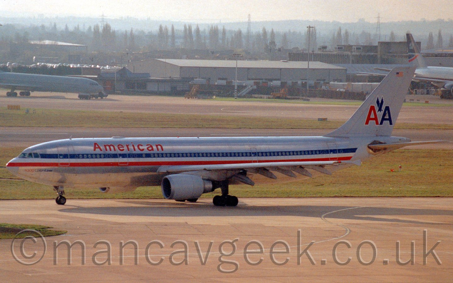 Side view of a twin engined jet airliner taxiing from right to left. The planes body is in a natural metal finish, with a blue, white, and red stripe running from nose to tail, and red "American" titles on the upper forward fuselage. There are a pair of capital letter "A"s, one red, the other blue, on the tail, with an image of a stpooping eagle between them. In the background, various large building line the airfield, with a pair of planes, in front of them, one plane on each side of the frame, both facing to the lower right corner. Threes are visible through the haze in the distance. The overall scene is a bit hazy and grainy, but with some strong sunlight from the left, with bright speckles on this planes forward fuselage, and a strong shadow on the ground.