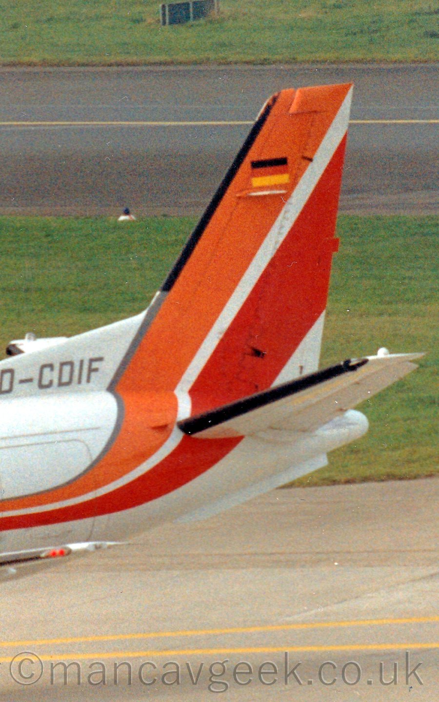 Closeup of the tail of a propellor-engined airliner taxiing from right to left. The plane is mostly white, with an orange and red stripe running along the body and sweeping up in to the tail. The registration "D-CDIF" is on the front of the base of the tail. In the background, a runway has grassed areas on either side.