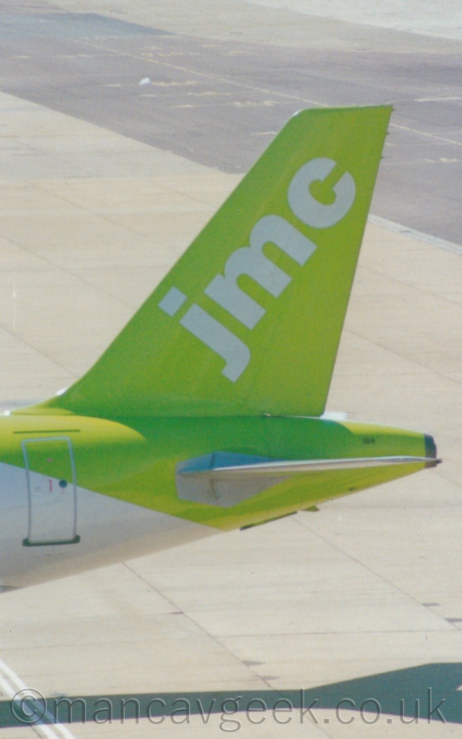 Closeup of the tail of a jet airliner parked facing to the left. The plane's body is mostly white, with a neon green rear fuselage and tail, with diagonal white "JMC" titles on the tail. In the background, acres of tarmac apron space fill the rest of the frame.