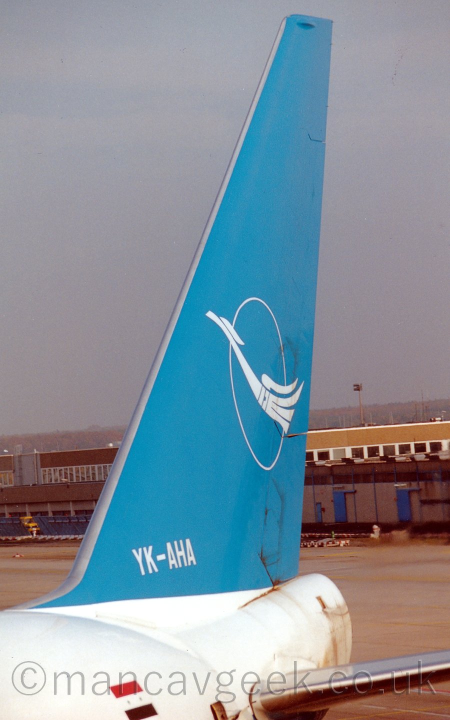 Closeup of the tail of a very large jet airliner parked on it's gate, facing to the left and towards the camera. The plane has a white body, with a sky blue tail carrying the image of a white bird flying past the outline of a white circle. The registration "YK-AHA" is on the base of the tail, painted in white. In the background, airport buildings fill the lower half of the image, with grey sky filling the rest.