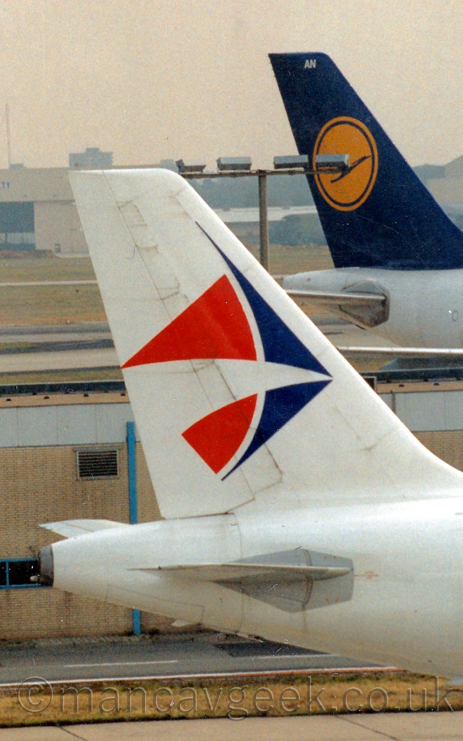 Closeup of the tail of a twin engined jet airliner taxiing from left to right. The plane is mostly white, with a blue and red logo on the tail that is somewhere between a child's kite, and an angel fish. In the background, a low, sandy coloured airport building can be seen, with the back end of a jet airlinervwith a white body and a blue and yellow tail just beyond that. In the far distance, a large white hangar with the doors partially open in the middle, and the words "safety - security" on the upper section, is slowly dissapearing into haze, under a hazy grey sky.