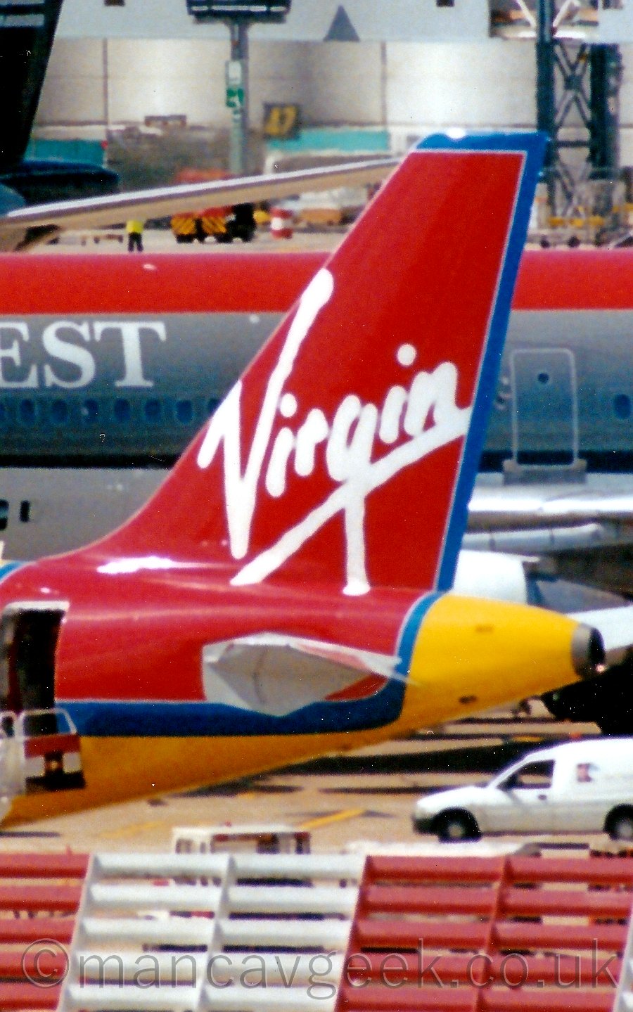 In the background, a large slatted metal barrier painted in alternating red and white sections fills the bottom of the screen, through which some vehicles can be see clustered around this plane. In the background, a red, grey, and white plane fills most of the rest of the frame, with a grey airport building in the distance filling what is left.