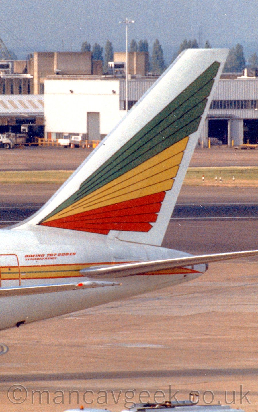 Close up of the tail and rear fuselage of a jet airliner taxiing to the left. The plane is mostly white, with a hellow stripe outlined in red running along the body, and green, yellow, and red stylised wing feathers on the white tail. Thewre are "Boeing 7676-200ER Extended Range" titles on the upper rear fuselage. In the background, a large white building takes up a good chunk of the frame, especially on the right, with another brown buiding behind it and to the left., with grey-blue skies above.
