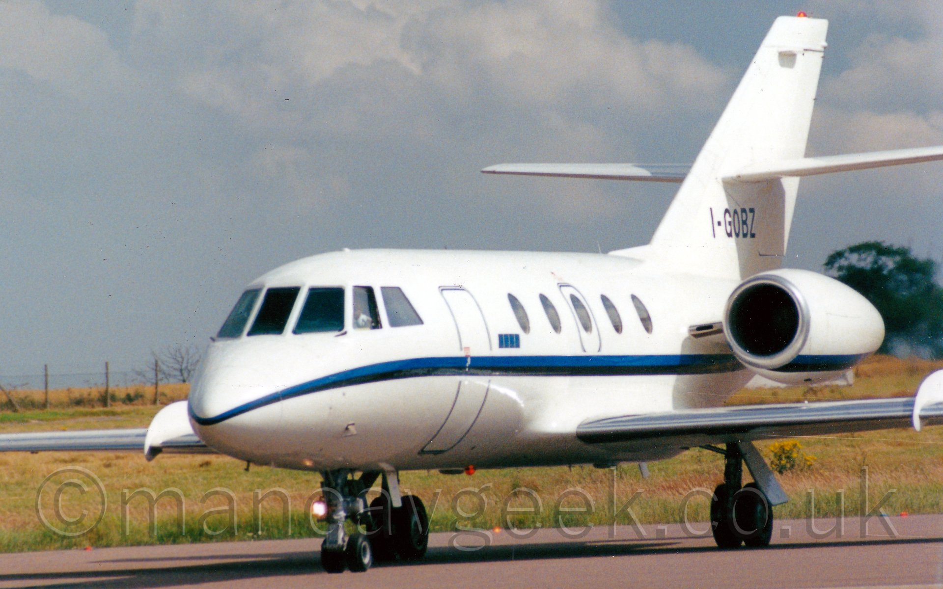 Side view of a twin engined bizjet parked facing to the left, and slightly towards the camera. The plane is almost entirely white, with a thin blue and black stripe running the length of the body, as well as along the bottom of the engine pods. The registration "I-GOBZ" is on the bottom half of the tail in black. In the background, grass runs off to a wire fence on the left of the frame, and trees on the right, under a wispy grey sky with patches of blue threatening to peek through.