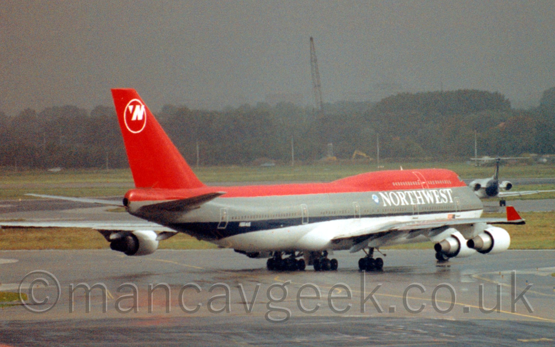 Side view of a very large 4 engined jet airliner taxiing from left to right and slightly away from the camera at a rather misty airport. The plane has a red top and light grey belly, with a thick dark grey stripe and thin black stripe running along the body, the black stripe getting wider towards the rear. There are white "Northwest" titles on the grey stripe on the upper forward fuselage, under the passenger cabin windows on the upper deck. The red tail has the white outkine of a circle at the top, with a large letter "N" in the top right, and a small white triangle pointing North-West in the top left. The upturned wingtips are also painted red, while the engine pods are painted white. In the background, a smaller, twin engined jet airliner is taxiing away from the camera on the right of the frame, with grass leading off to trees and a dismal grey sky filling the rest of the frame.