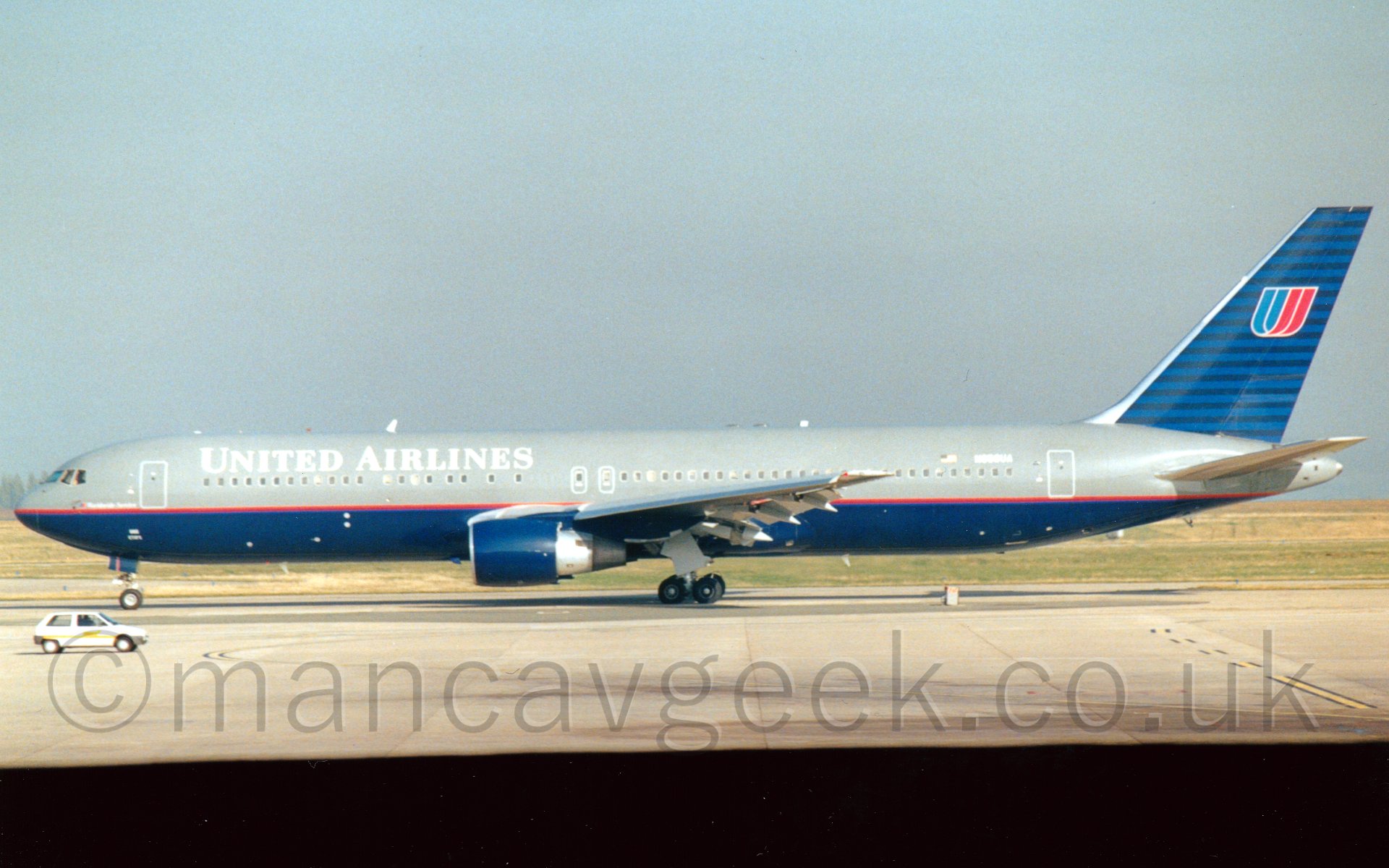 Side view of a twin engined jet airliner taxiing from right to left. The plane is mostly grey, with a dark blue belly, and a thin red stripe running along the body, seperating the 2 colours. There are white "United Airlines" t4itles on the upper forward fuselage, and the registration "N656UA" on the upper rear fuselage, also in white. The engine pods are the same dark blue, while the tail has a series of thin horizontal stripes in dark and mid-blue, with a lette "U", seemingly formed from a twisted ribbon, one side red, the other light blue. In the foreground, a small white car is on the lower left of the frame, driving to the right past the plane's nosewheel. In the background, grass runs off to the horizon, under a flat grey sky.