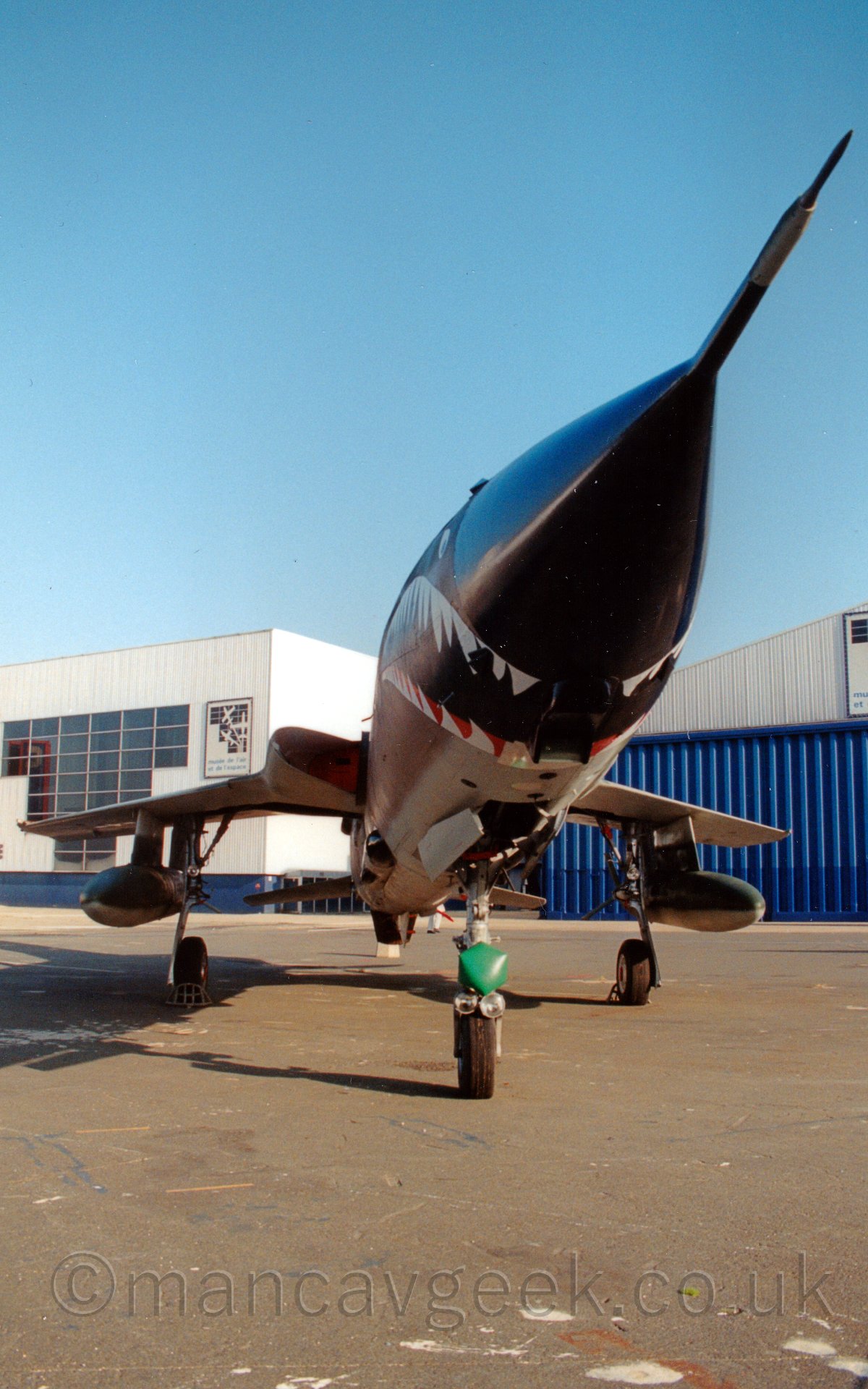 Front view from below and slightly to the left of a single-engined, single-seat jet fighter, parked facing slightly to the right of the camera. The plane is mostly painted a muddy brown, with a light grey belly, and a sharks mouth painted on the lower forward fuselage, behind the black nosecone, which itself has a large probe poking forward, towards the top right corner of the frame. There are large drop tanks suspended below the wings, and a large green light on the nosewheel door. In the background, a large white building stretches across the frame, the bit on the right having large blue hangar doors. Above, the sky is a gorgeous blue.
