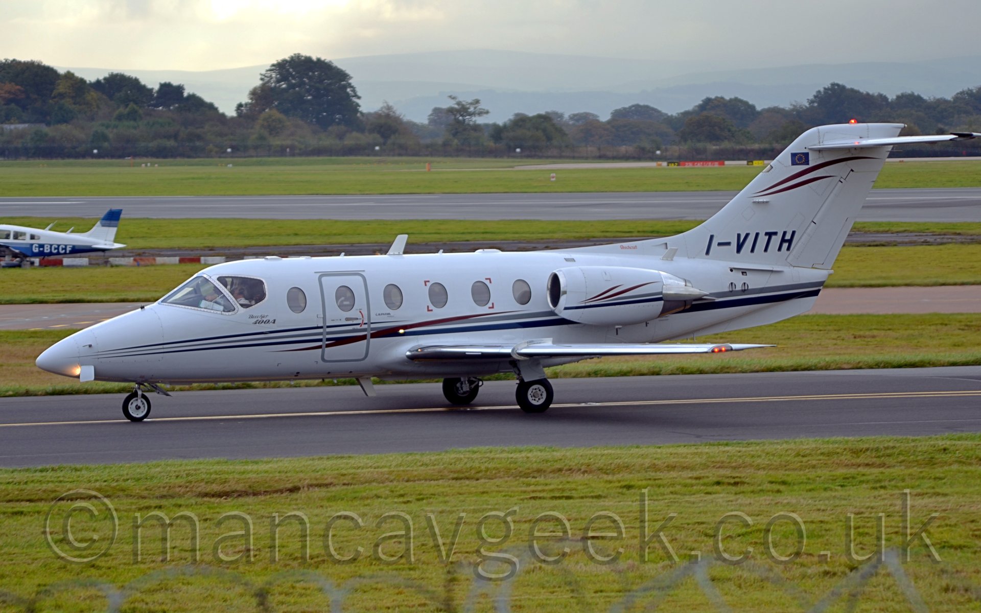 Side view of a twin engined bizjet taxiing from right to left. The plane is mostly white, with blue, grey, and red stripes running along the body and along the engines. The registration "I-VITH" is on the base of the tail in black, under more red stripes. Grass fills the lower part of the frame, with more grass, taxiways, and a runway leading off to trees in the distance. There is a single-engined blue and white light aircraft poking it's tail in to the middle left of the frame. Rolling hills in the far distance slowly fade into the hazy white sky.