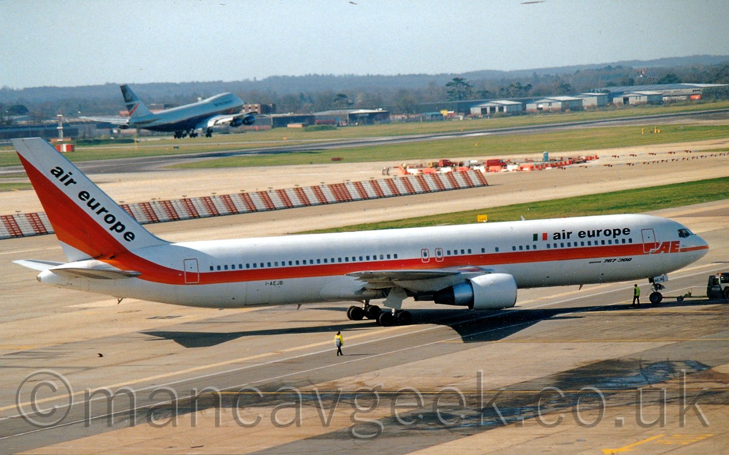 Side view of a twin engined jet airliner being pushed back from it's gate, to the left, by a grey tug attached to the nosewheel, with someone in a hi-viz vest walking alongside the nosewheel, and another person in a hi-viz jacket walking by the wingtip. The plane is mostly white, with a red and orange stripe running along the body and sweeping up in to the tail. There are black "air europe" titles on the upper forward fuselage, next to an Italian flag (vertical green, white, and orange bars), and diagonally on the tail. In the background, a white and red striped metal barrier runs most of the way across the frame from the left, seperating 2 sections of taxiway, with a small construction site at the end. In the background, a runway runs from one side of the frame to the other, lined with buildings on the far side, and with a very large dark blue and grey plane coming in to land, flying just feet above it. Trees lead off in to the distance, meeting a bright but hazy sky.