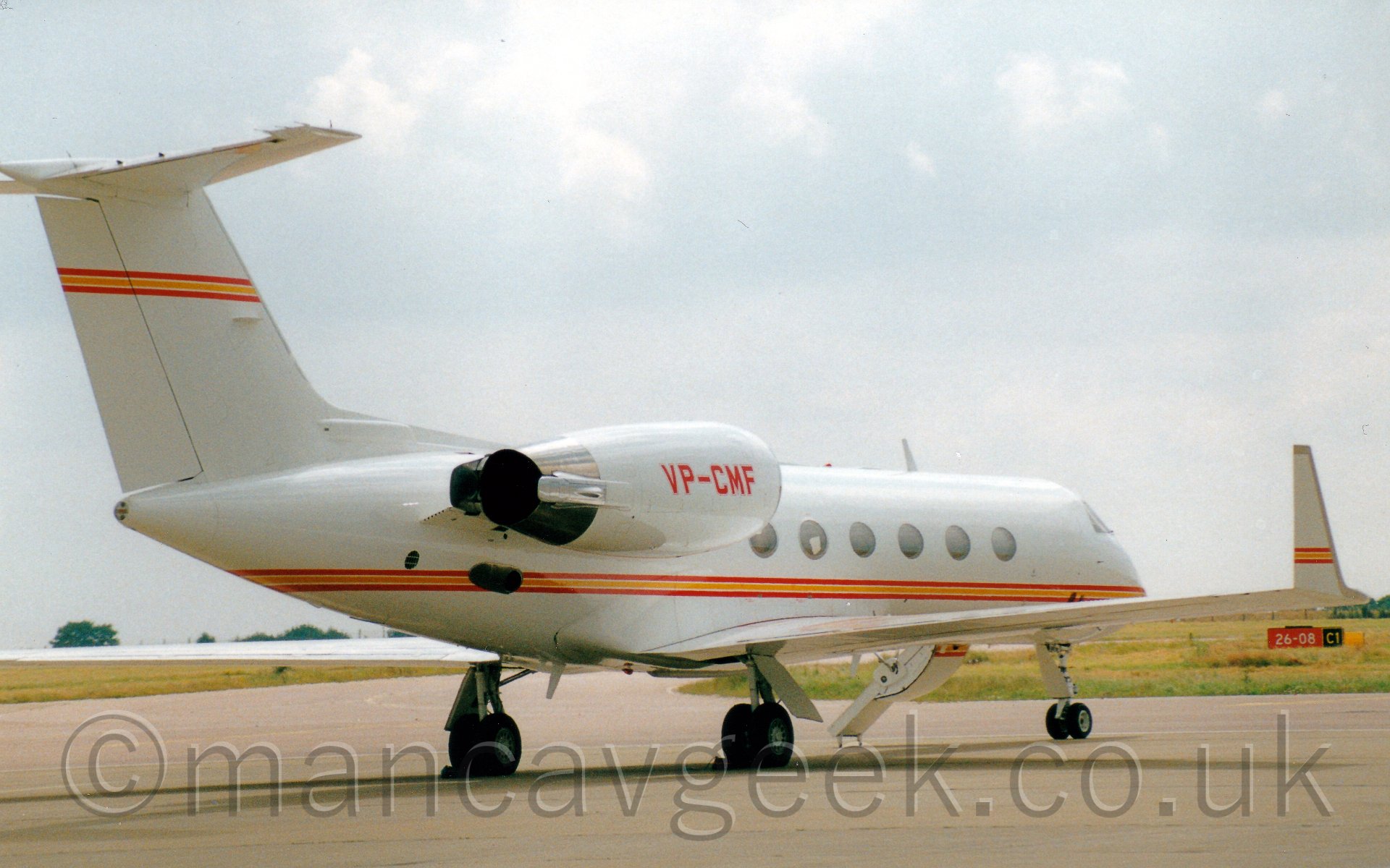 Rear side view of a twin engined bizjet parked facing to the right and away from the camera. The plane is mostly white, with a red and orange stripe running along the body, as well as along the tail and the upturned wingtips. The registration "VP-CMF" is on the engine pods in red. There is a door open on thefar side of the plane, visible under the fuselage, with it's built-inairstairs extended. In the background, grass leads off to trees on the horizon, under a bright white sky.