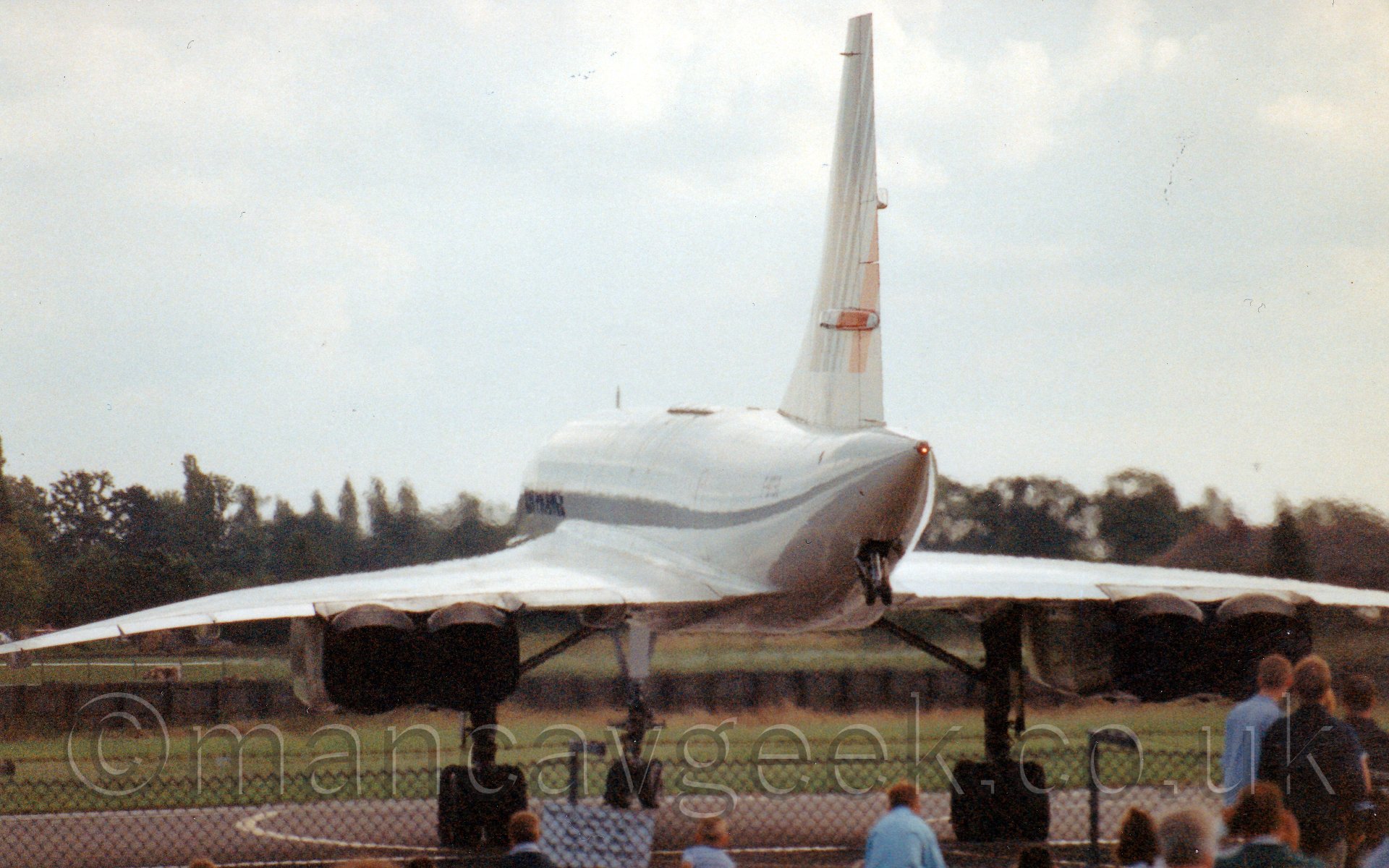 Rear view of a 4 engined, very thin, very long, very pointy jet airliner, with wings tilted slightly down, facing almost directly away from the camera and turning to the right. The plane is almost entirely white, with a series of red and blue diagonal stripes on the tail. A set of wheels, not connected to the ground, can be seen coming from below the tailcone. In the foreground, a group of people can be seen watching it through a chain link fence topped with barbed wire, while in the background, grass and trees meet a bright white sky.