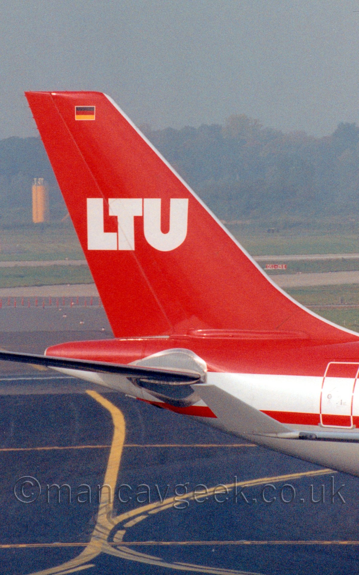 Closeup of the tail of a twin engined jet airliner taxiing from left to right. The plane has ared upper section and a grey belly, with a white and red stripe running along the middle of the fuselage. The tail is red, and has white letters "LTU" emblazoned across the middle, with a German flag (equal black, red, and yellow horizontal bands" outlined in white at the top. The planes grey upturned right wingtip is visible towards the middle of the frame. In the background, haze renders the large grassed areas and trees at the perimeter almost indistinguishable, under a dismal grey sky.