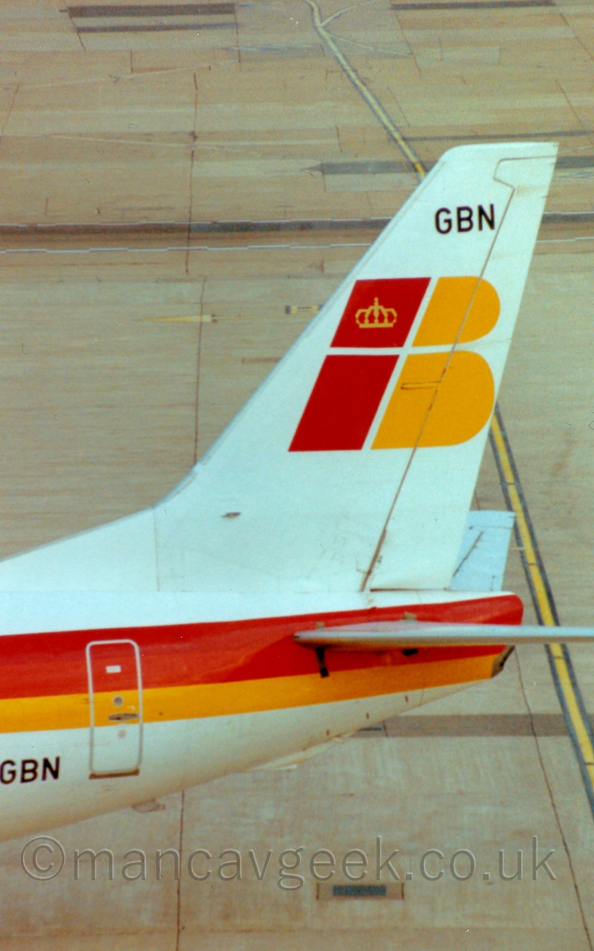 Closeup of the tail of a jet airliner taxiing from right to left. The plane is mostly white, with a thick red and yellow stripe running along the body. The tail is also white, with large red lower case letters "IB" in red and yellow respectively, the "I" having a golden crown in the dot at the top. Smaller letters "GBN" can be seen in black at the top of the tail, and on the lower rear fuselage. The background is entirely made up of concrete slabs of apron area.