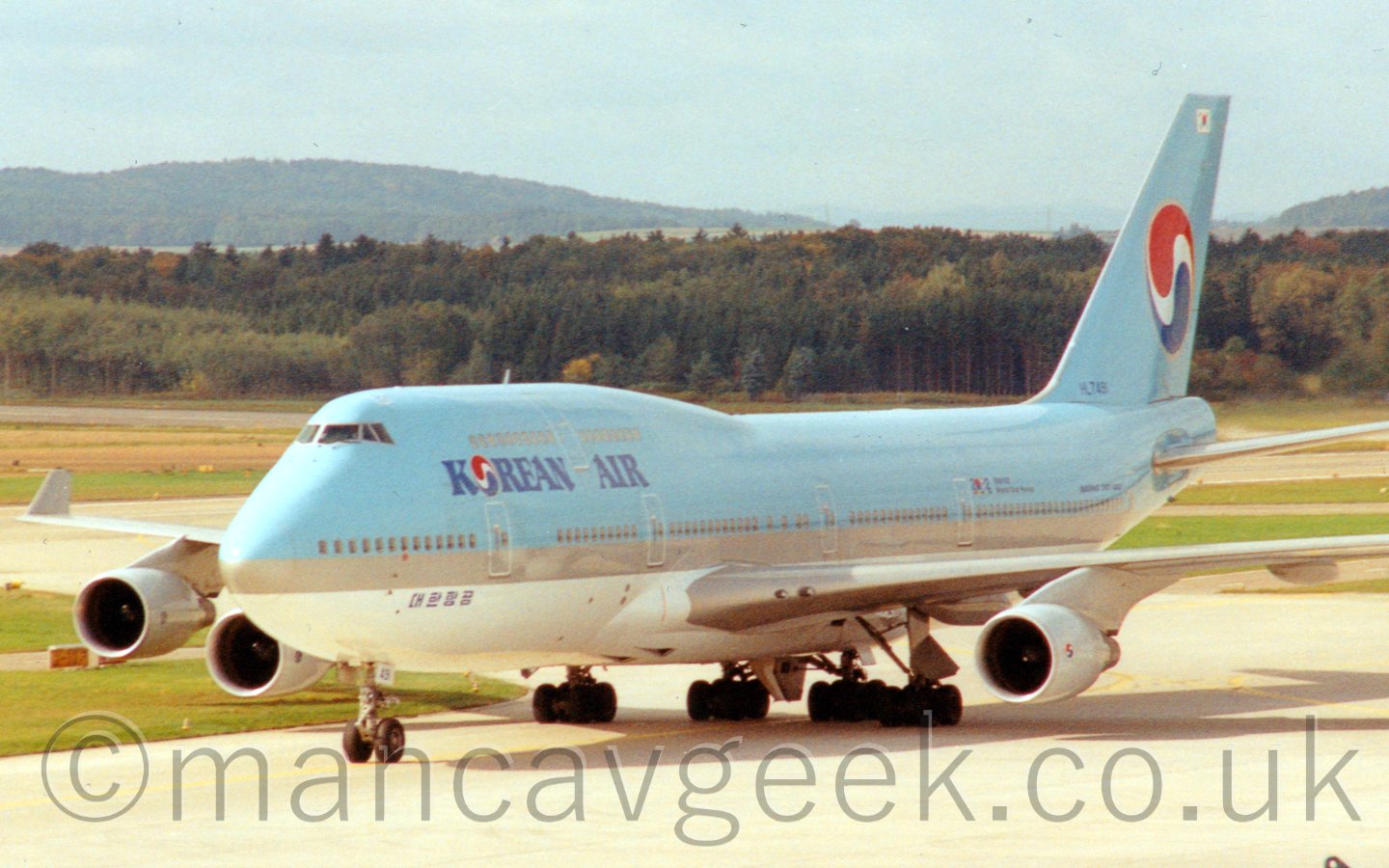 Side view of a blue, very large, 4 engined jet airliner taxiing from right to left and slightly towards the camera. The plane has a white belly, and blue Korean Air" titles on the upper forward fuselage, with a red, white, and blue Yin-Yang symbol on the tail. In the background, tree-covered rolling hills meet the hazy blue sky.