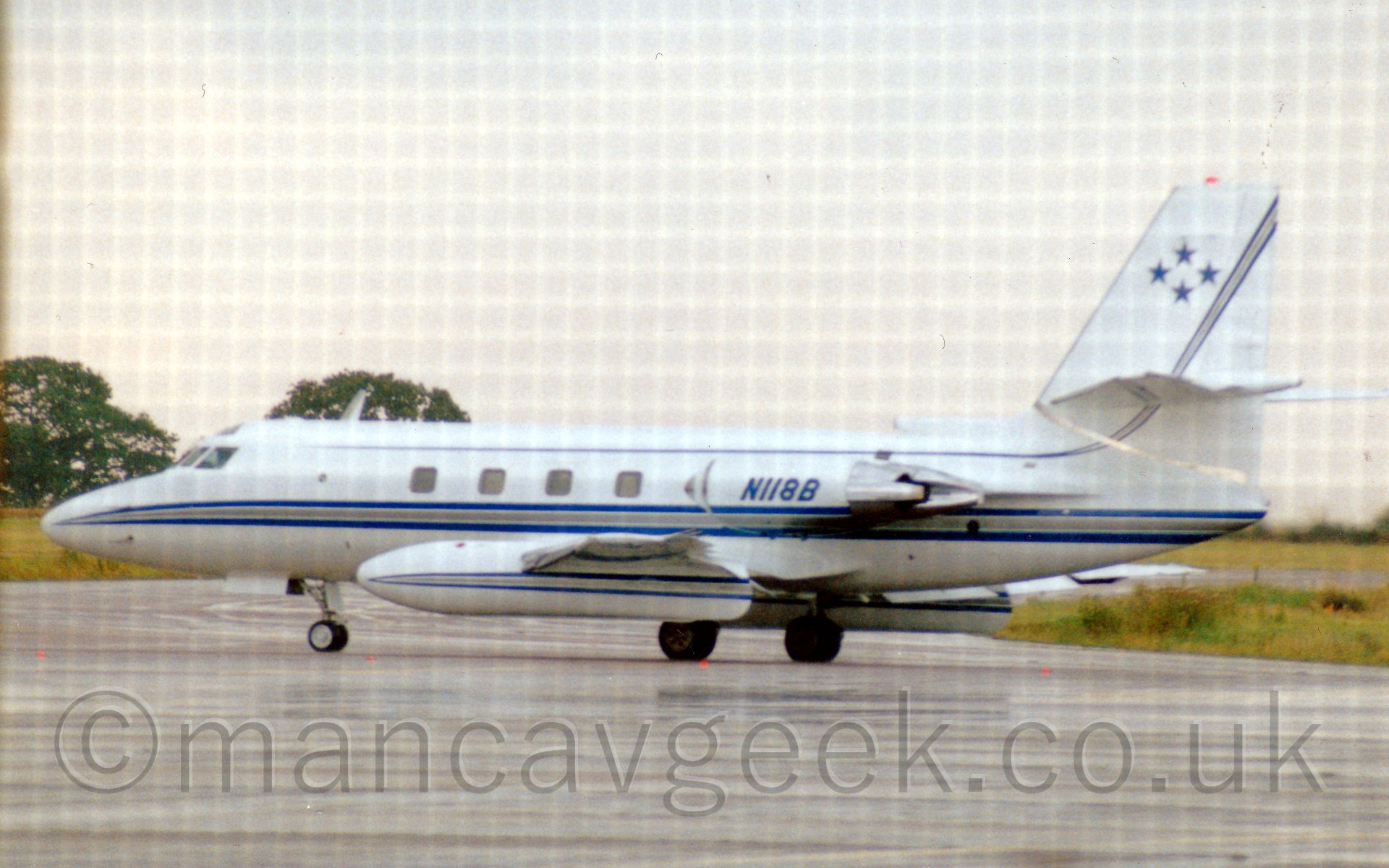 Side view of a 4 engined bizjet, with the engines mounted in pairs on either side of the rear fuselage, taxiing from right to left. The plane is mostly white, with a blue and grey cheatline running along the fuselage and lower parts of the engine pods, as well as along the wing-mounted external fueltanks. A second, thinner, blue cheatline runs along the top of the fuselage, sweeping up into the tail where it widens out to reveal the grey middle. The registration "N118B" is on the engione pods in blue, while the tail has 4 5 pointed blue stars arranged in a diamond pattern. In the background, grass runs off into the distance, with trees visible on the horizon on the left of the frame, under a bright but hazy sky. A fine, square moire pattern covers the entire image, probably caused by shooting through a fine mesh fence.