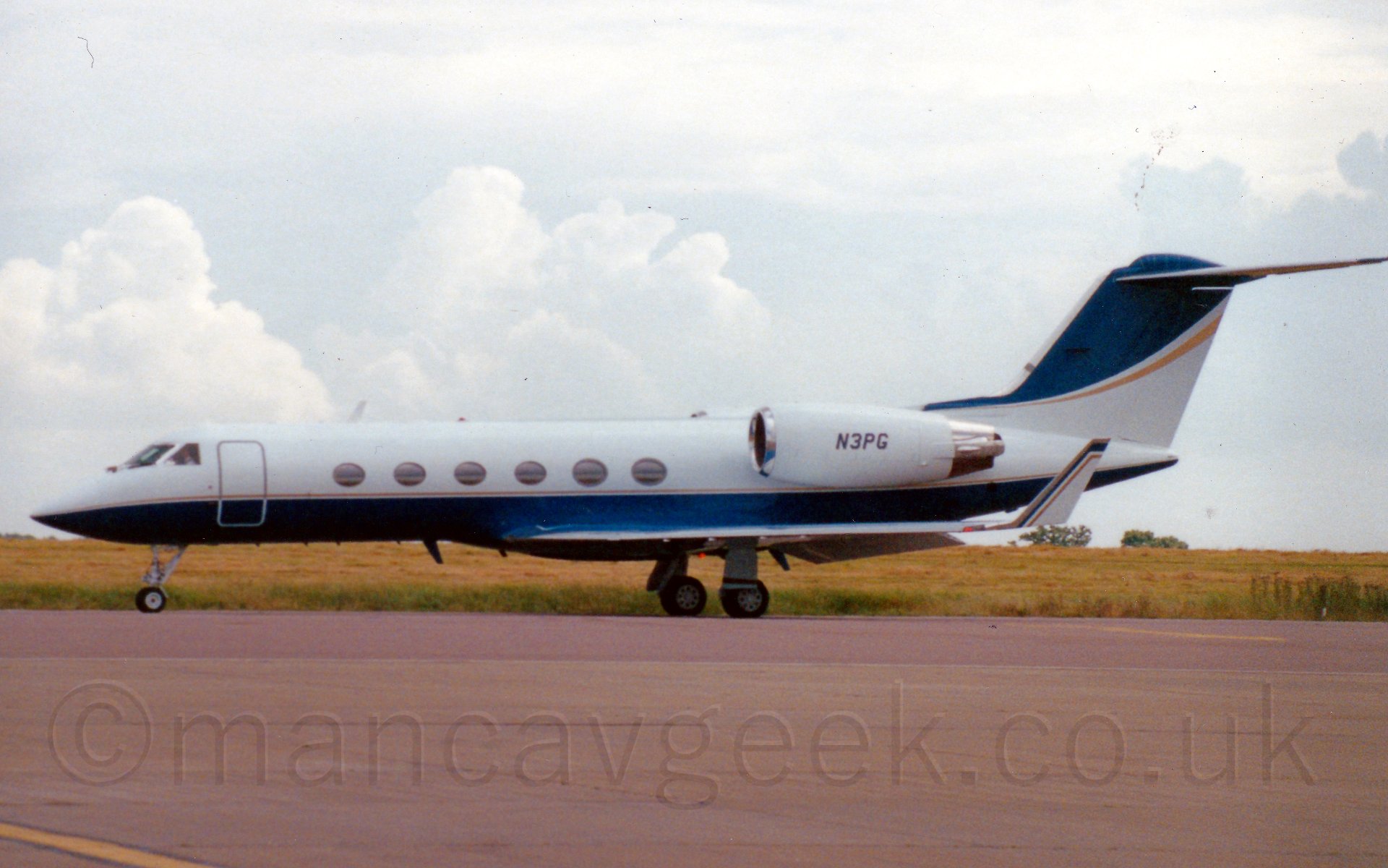 Side view of a twin engined bizjet parked facing to the left. The plane is mostly white, with a blue belly, and a thin golden cheatline. The tail and upturned wingtips have the colours inverted, blue at the front and top, white at the bottom and rear, with a golden line separating them. The registration "N3PG" is on the white engine pods, in black. Tarmac fills the foreground, with grass in the background running off to the horizon, where it meets fluffy grey clouds.