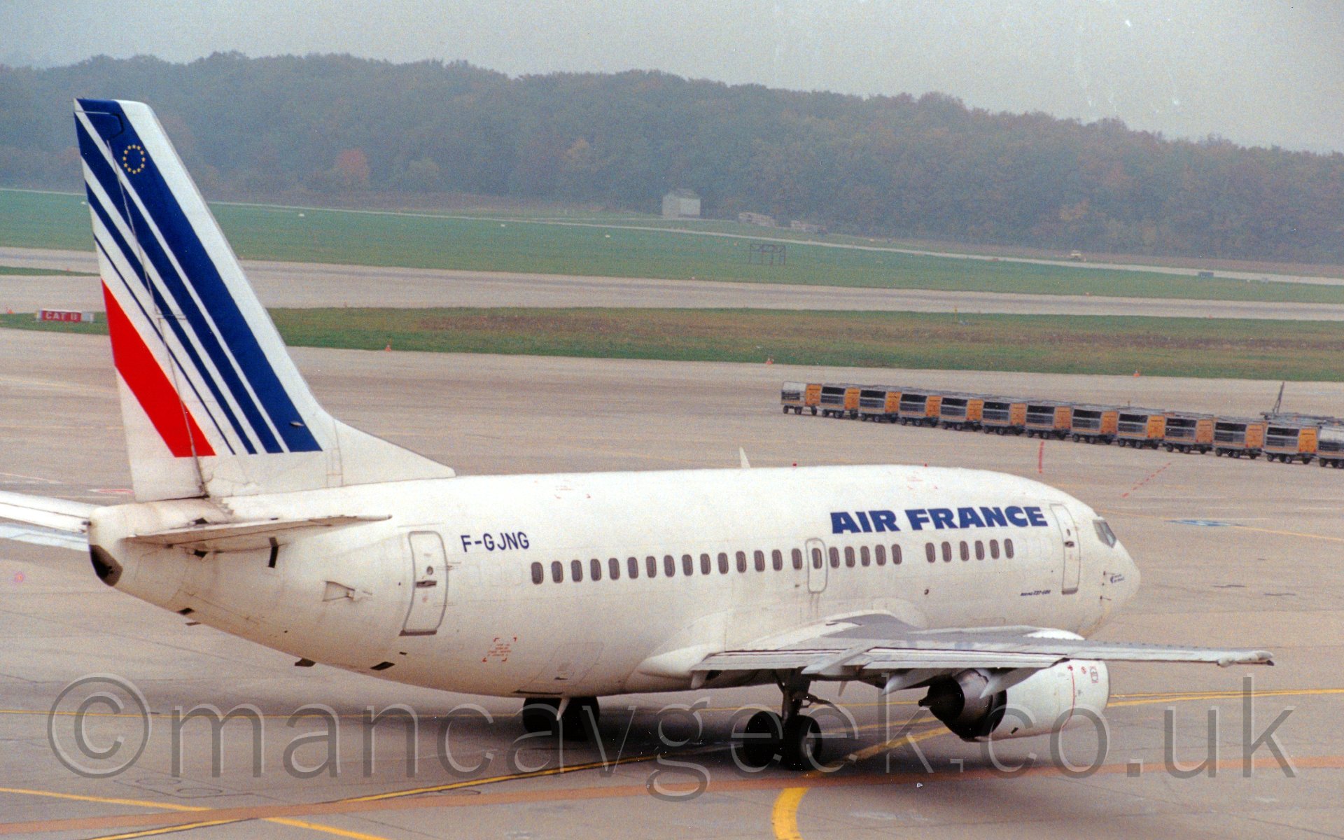 Side view of a twin engined jet airliner taxiing from left to rigtht and slightly away from the camera. The plane is mostly white, with blue "Air France" titles on the upper forward fuselage, and the registration "F-GJNG" on the upper reaer fuselage. The tail is white, with a series of diagonal blue stripes, getting thinner as they move to the rear, and a single red stripe. In the backgrounhd, a line of grey and yellow lugage carts stretch across the middle right of the frame, with grey taxiways and grassed areas leading up to trees meeting the hazy grey sky in the distance.