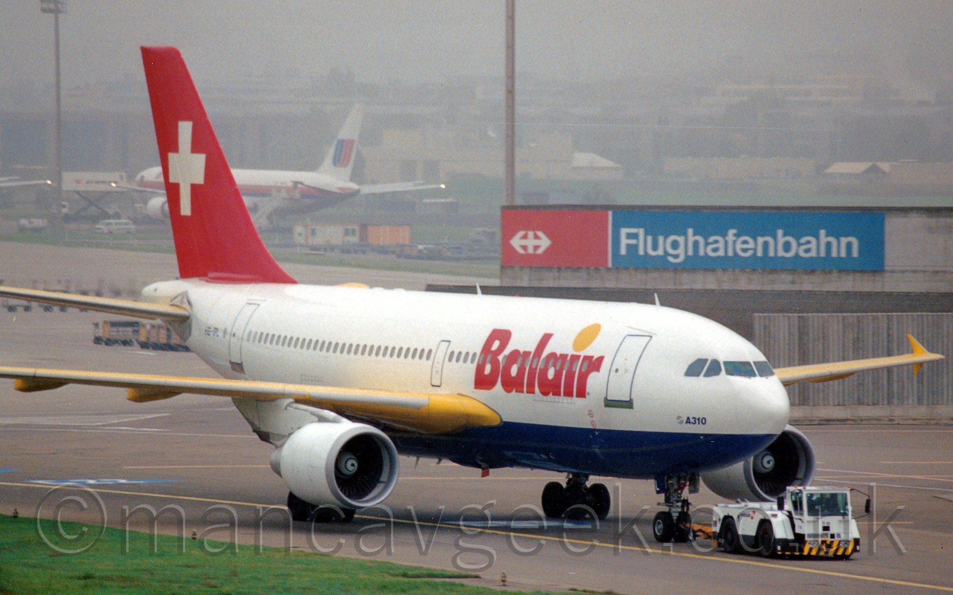 Side view of a twin engined white jet airliner with a blue belly and a red tail being towed from left to right by a white tug. The plane has red "Balair" titles on the lower forward fuselage, and a large white cross on the tail. In the In the background, a large blue and red sign with white "Flughafenbahn" titles adorns the side of a low building, with planes clustered around larger buildings, vanishing in to haze in the distance.