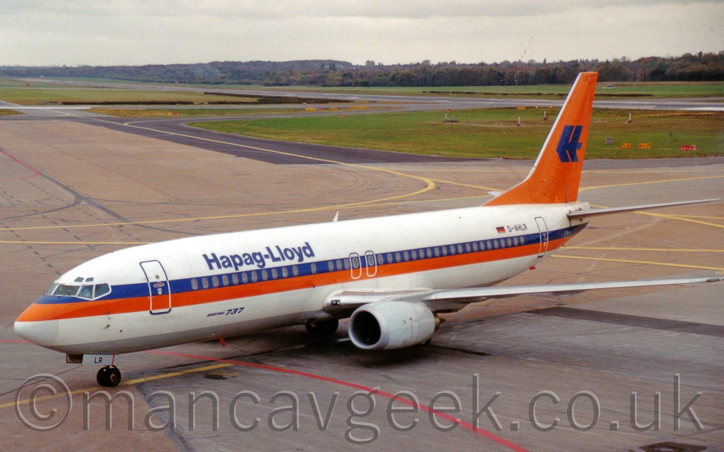 The plane is mostly white, with a grey belly, and an orange and blue cheatline running along the body, with blue "Hapag-Lloyd" titles on the upper forward fuselage. The tail is orange, with a rather angular capital letter "H" in the middle, looking similar to a double arrow head. In the background, taxiways give way to grass and a long runway, with more grass and trees beyond that, under a grey sky.