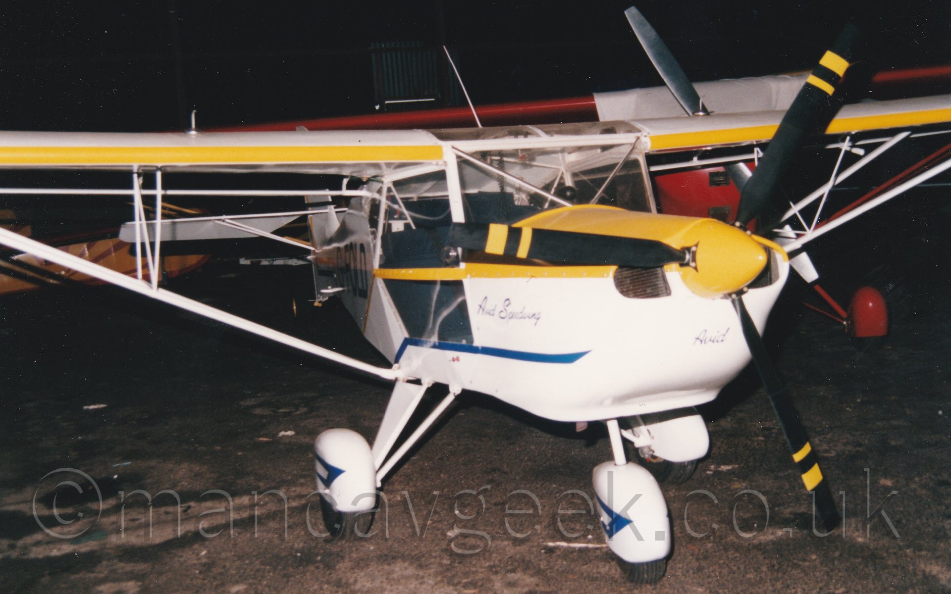 Front view of a high-winged, single seat light aircraft parked in a rather dim hangar. The plane is mostly white, with with a blue stripe running along the lower parts of the body and the wheel spats, and a yellow stripe running along the body from the yellow top engine cowlling panel. The registration "G-FOLD" is just discernable on the rear fuselage, with smaller blue "Avid" titles under the propellor, and "Avid Speedwing" on the side of the engine cowling, both in an italic font. IN the backb=ground, several other light aircraft can be seen in the murky depths of the hangar.